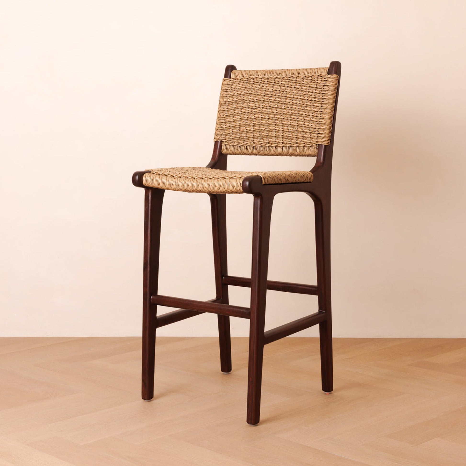 A wooden bar stool with a woven natural-fiber seat and backrest, shown on a light wood floor against a plain beige wall.