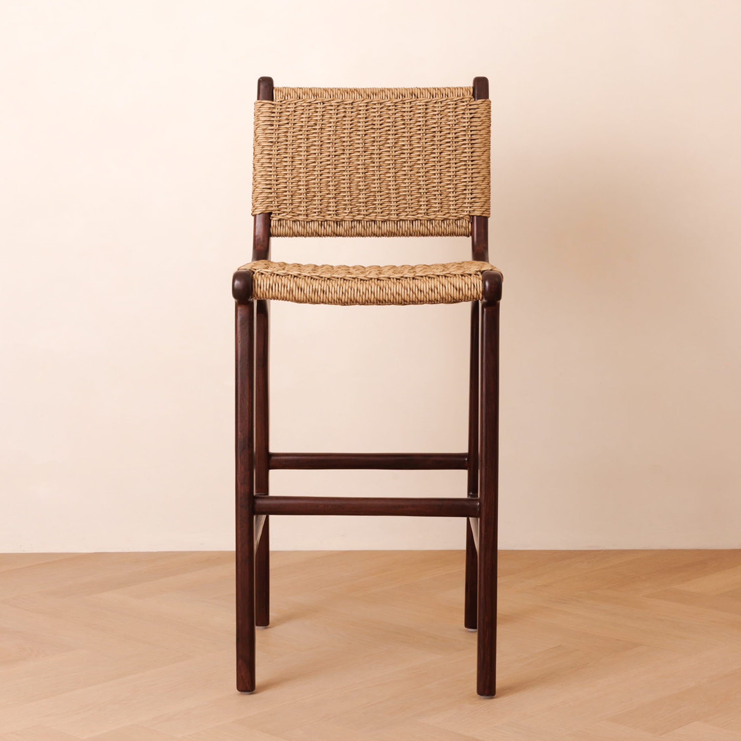 A wooden bar stool with a dark brown frame and natural woven jute rope seat and backrest, set against a minimalist beige wall and light wood floor.