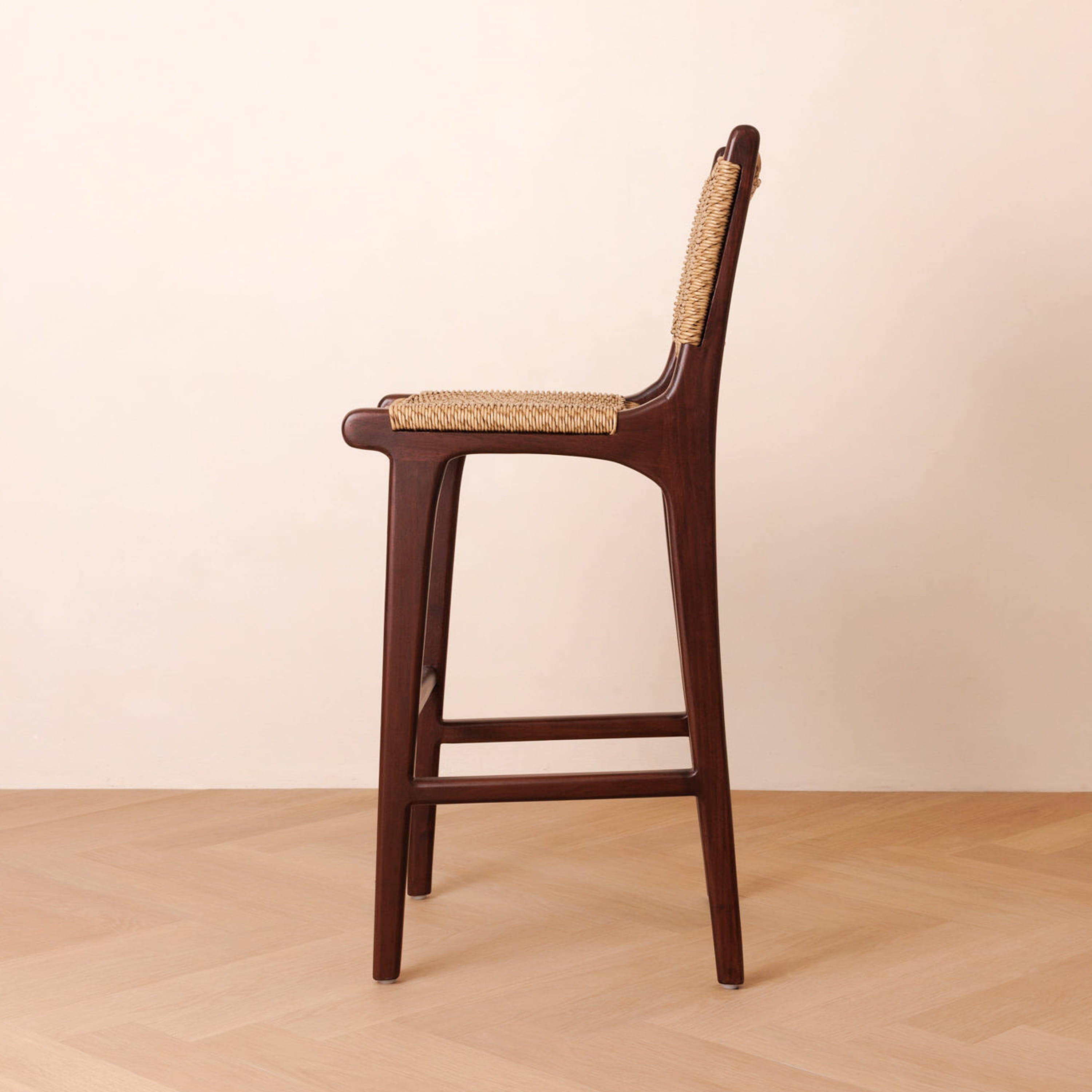 A single wooden bar stool shown from the side against a plain beige wall and light wood floor. The stool has a dark brown wood frame with a woven natural fiber seat and backrest.