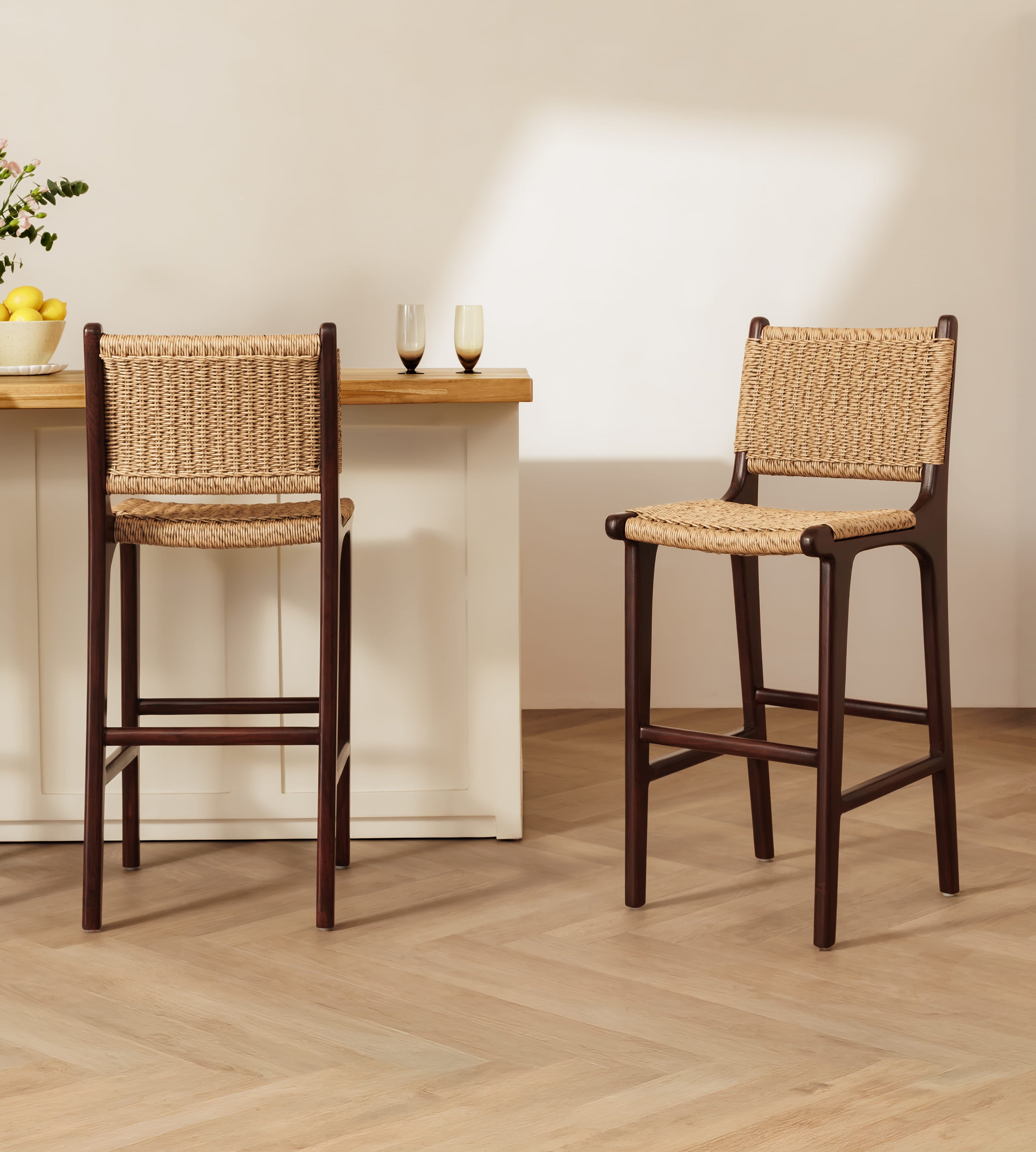 Two wooden bar stools with woven rope seats and backs, shown in a bright kitchen setting with a light wood countertop and herringbone floor.