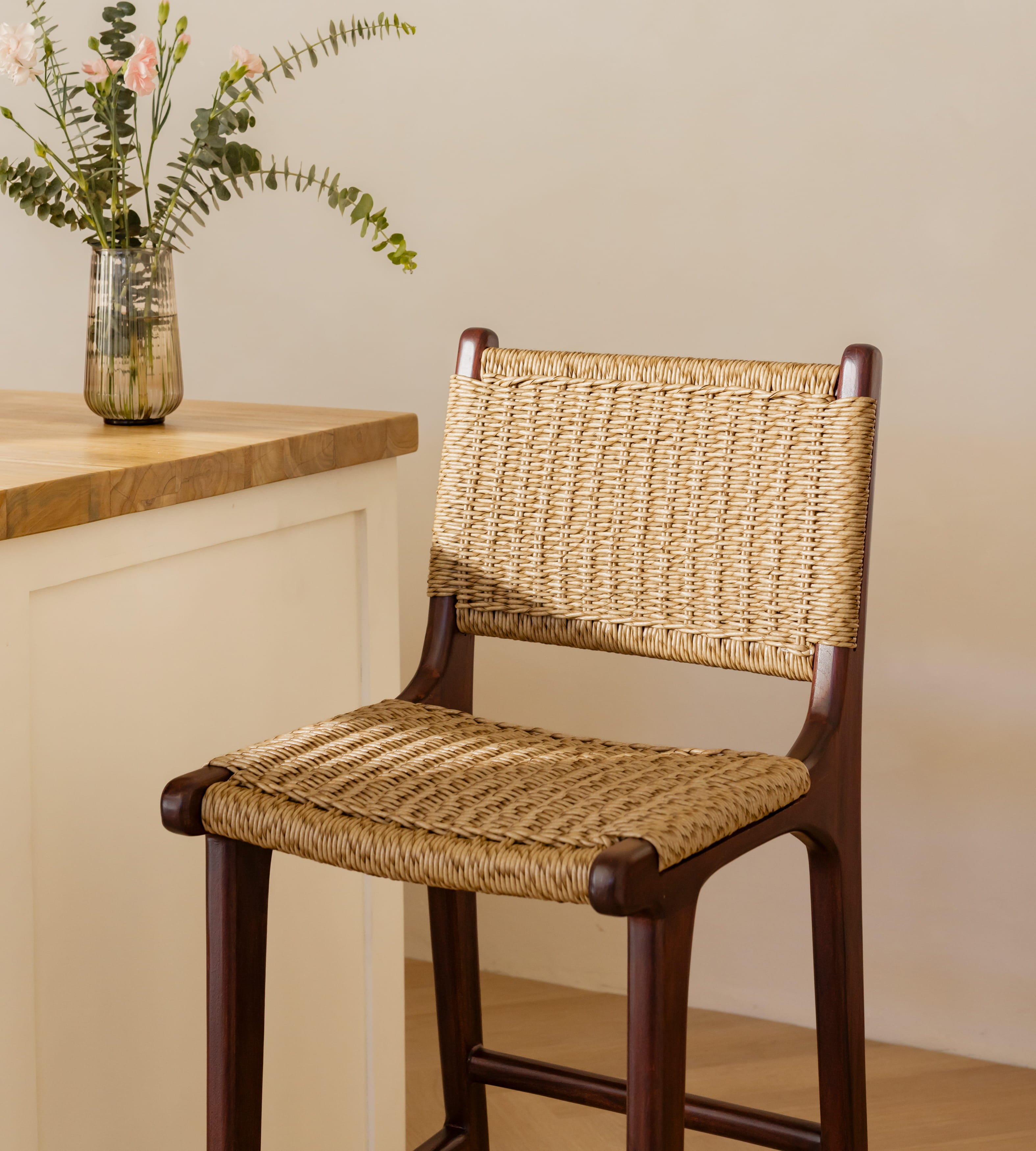A wooden bar stool with a woven natural fiber seat and backrest, positioned next to a light wood countertop with a glass vase of flowers.