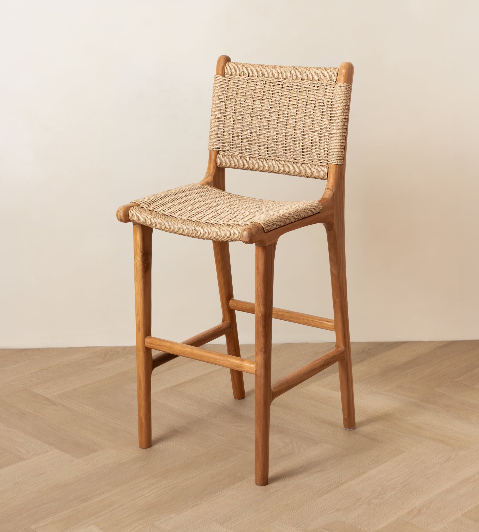 A wooden bar stool with a woven rattan seat and backrest, shown in a minimalist room setting with a light beige wall and wood-look floor.