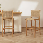 Two rattan bar stools with woven rattan seats and backs, shown in a bright, modern kitchen or dining setting with a light wood table and pale wall.