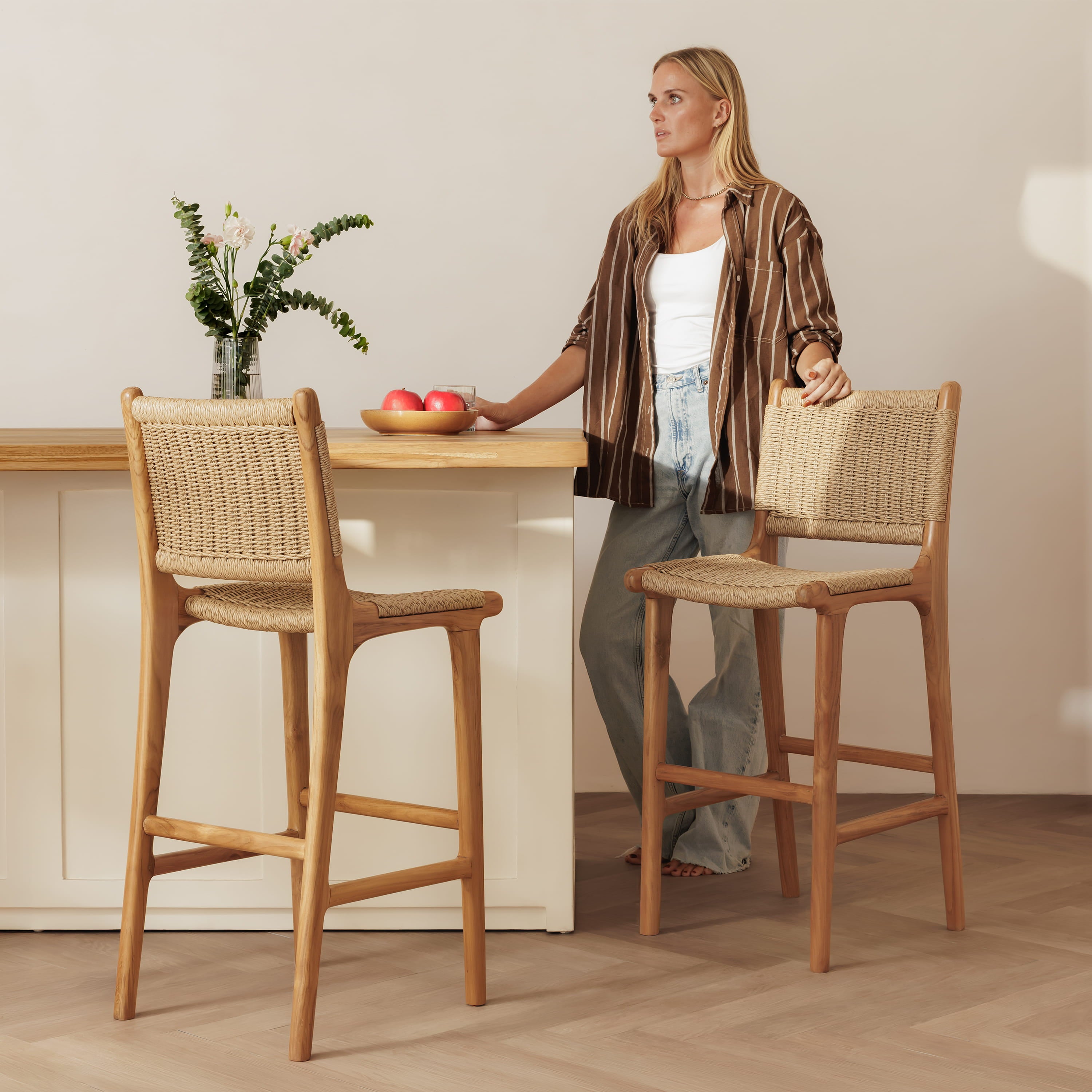 A woman stands beside a kitchen island holding the handles of two wooden bar stools with woven seats and backs, set in a bright, modern kitchen with light wood floors and a cream wall.