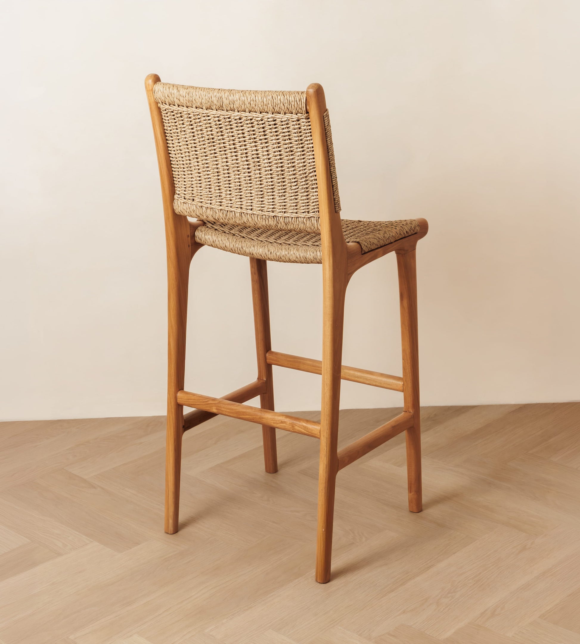 A wooden bar stool with a woven natural fiber seat and backrest, shown from a rear three-quarter angle on a light wood herringbone floor against a neutral beige wall.