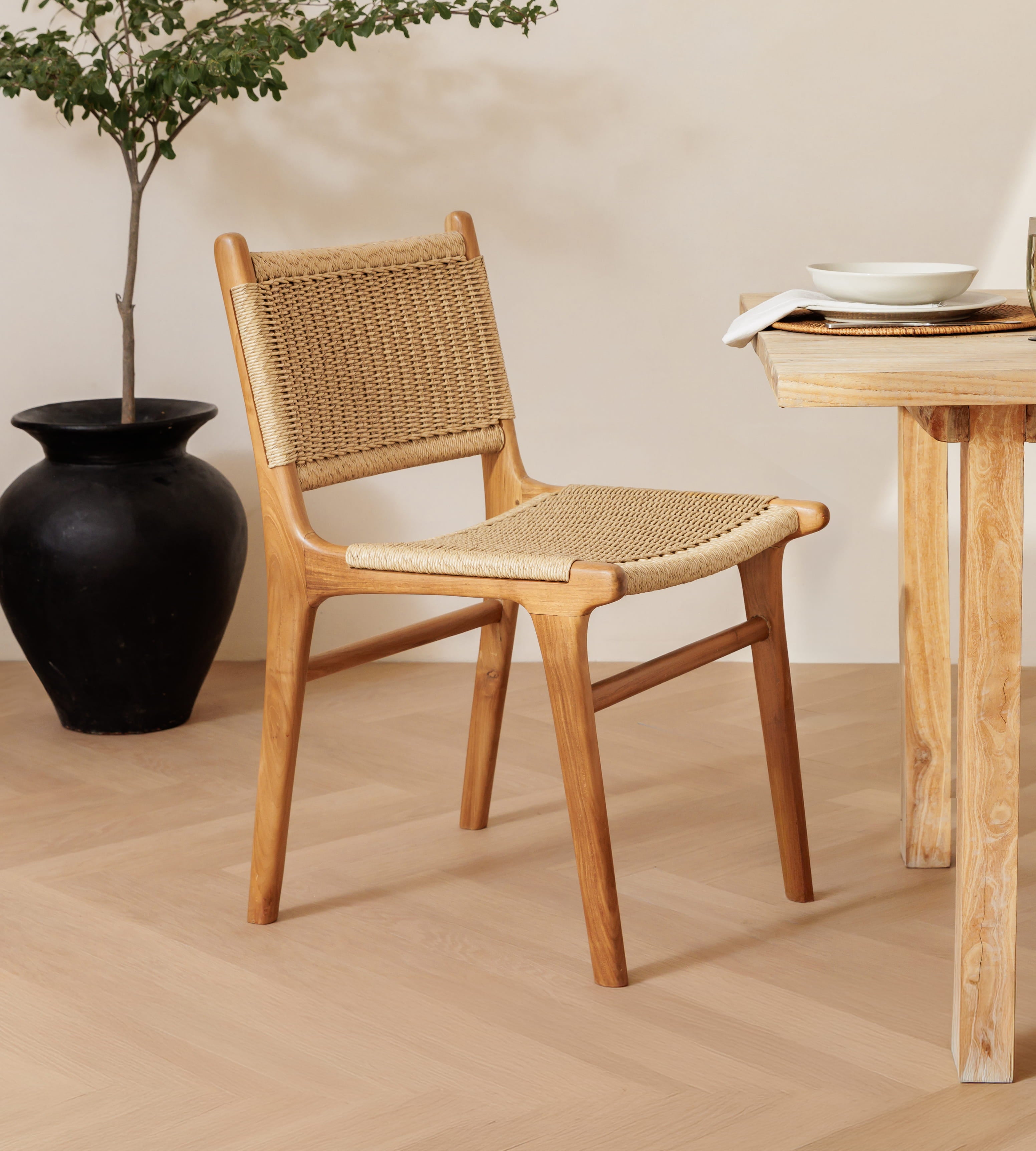 A rattan dining chair with a woven back and seat, positioned next to a matching wooden table in a warm, minimalist room with a black ceramic planter.