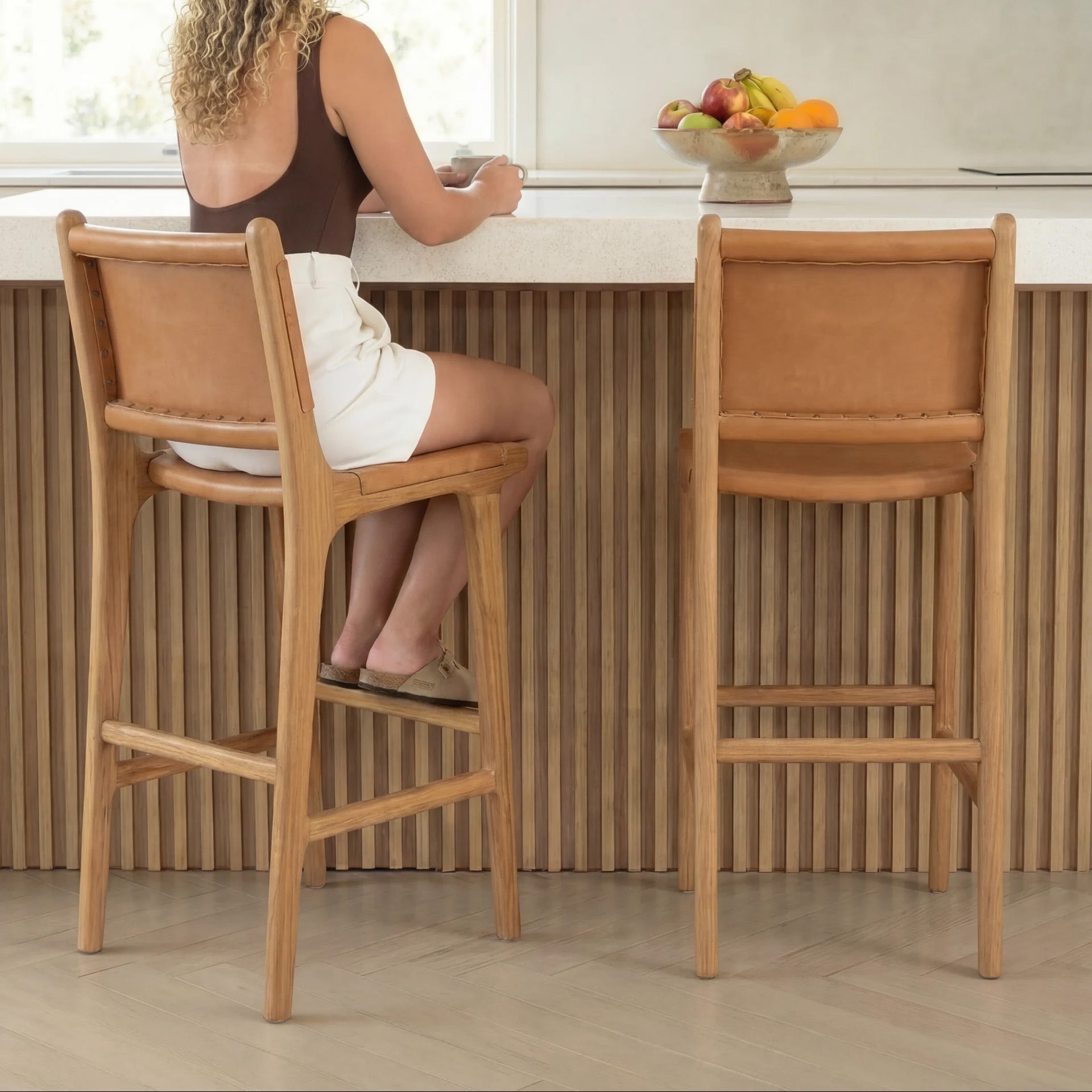 Woman sitting on a wooden bar stool in a kitchen, looking out a window.