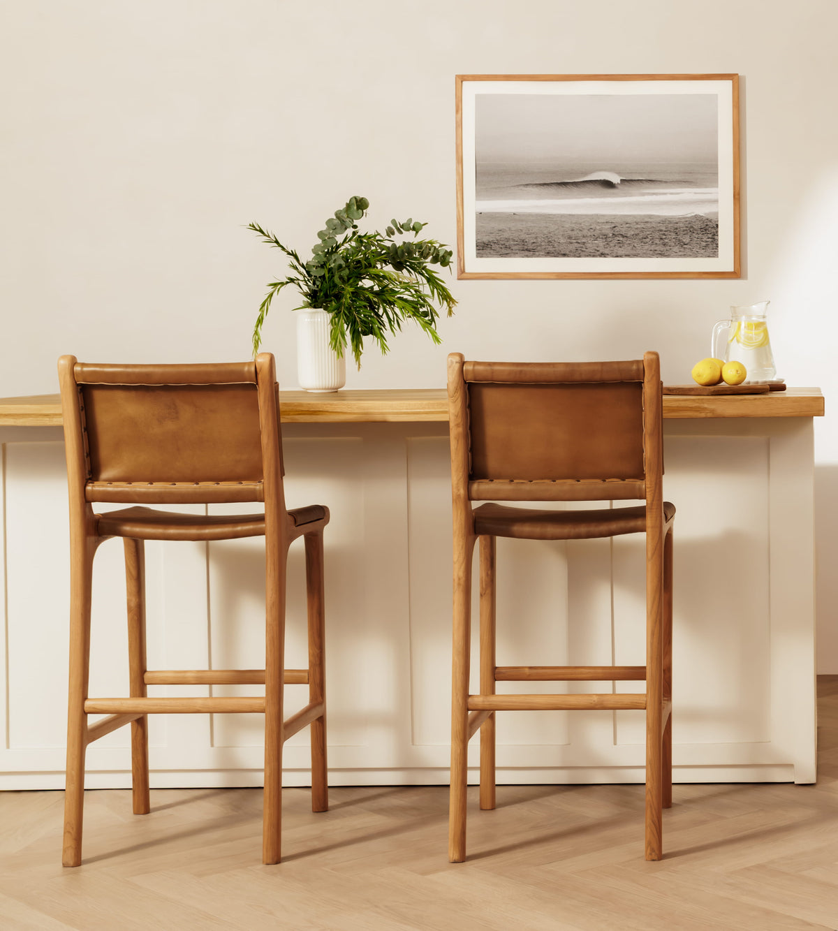 Two wooden bar stools with tan leather sling backs and seats, positioned in front of a white kitchen island with a framed black-and-white ocean print on the wall.