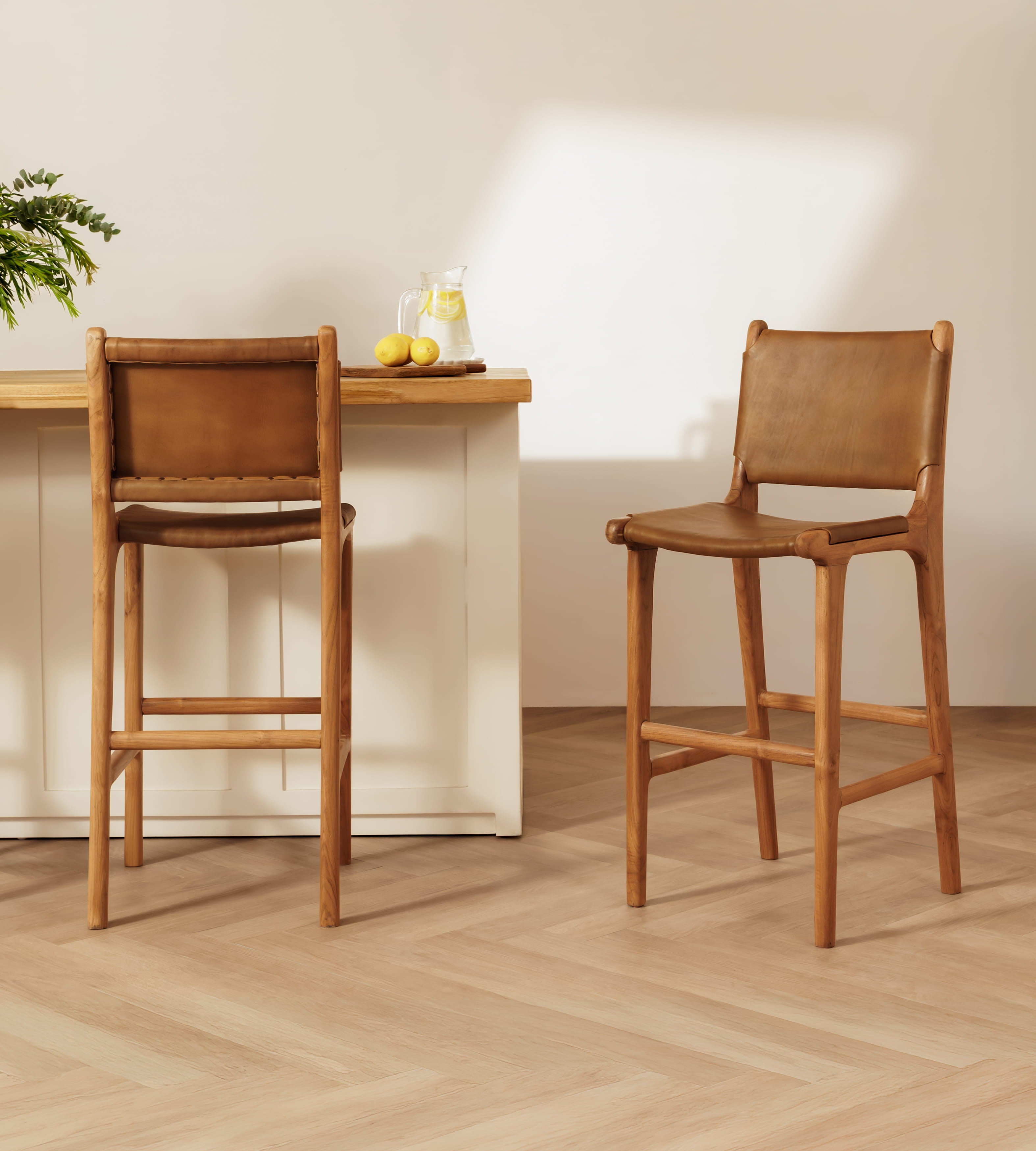 Two wooden bar stools with tan leather seats and backs, shown in a bright, modern kitchen setting with a light wood island and pale walls.