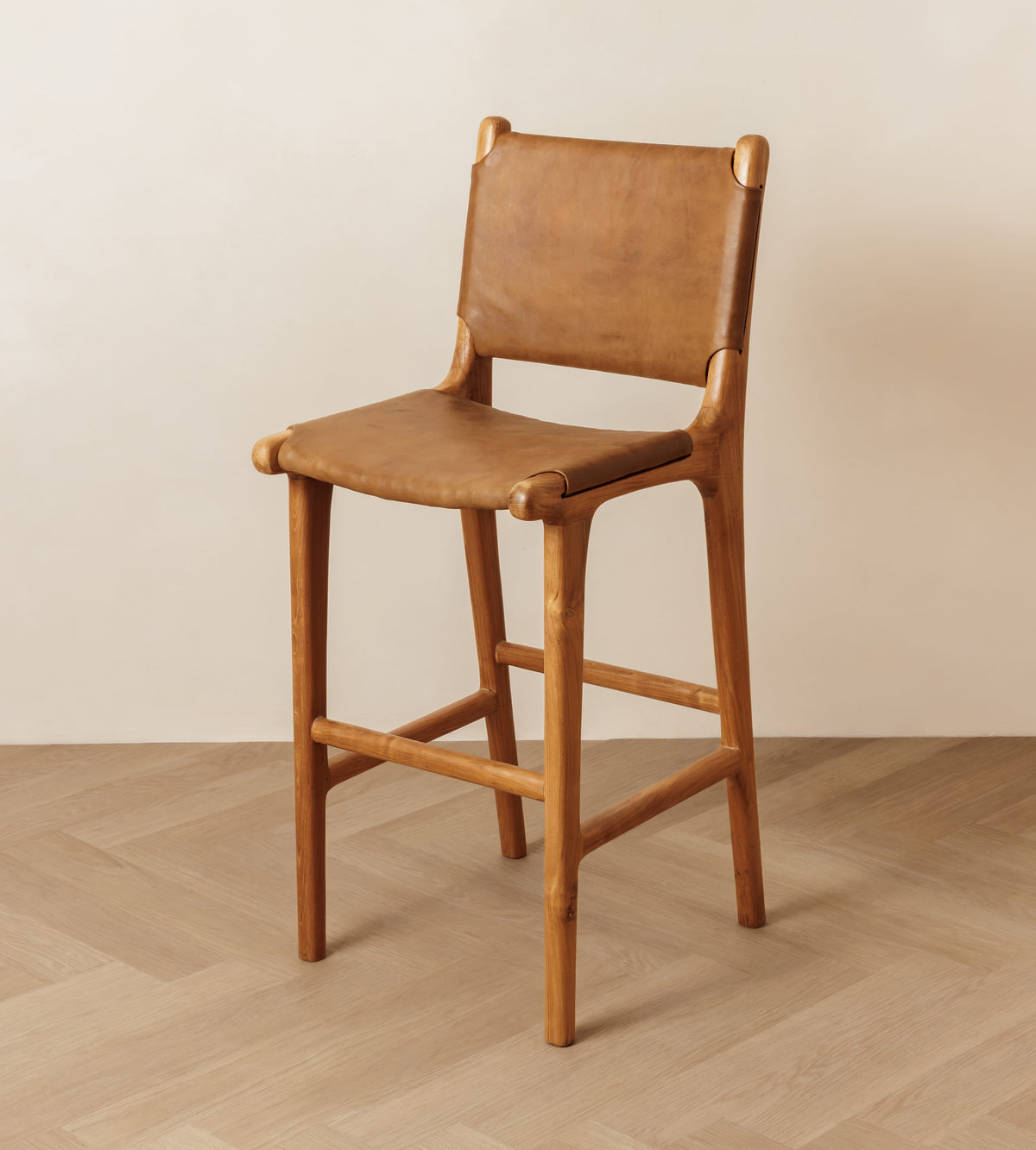 A mid-century modern wooden bar stool with a tan leather seat and backrest, set against a minimalist beige wall and light wood herringbone floor.