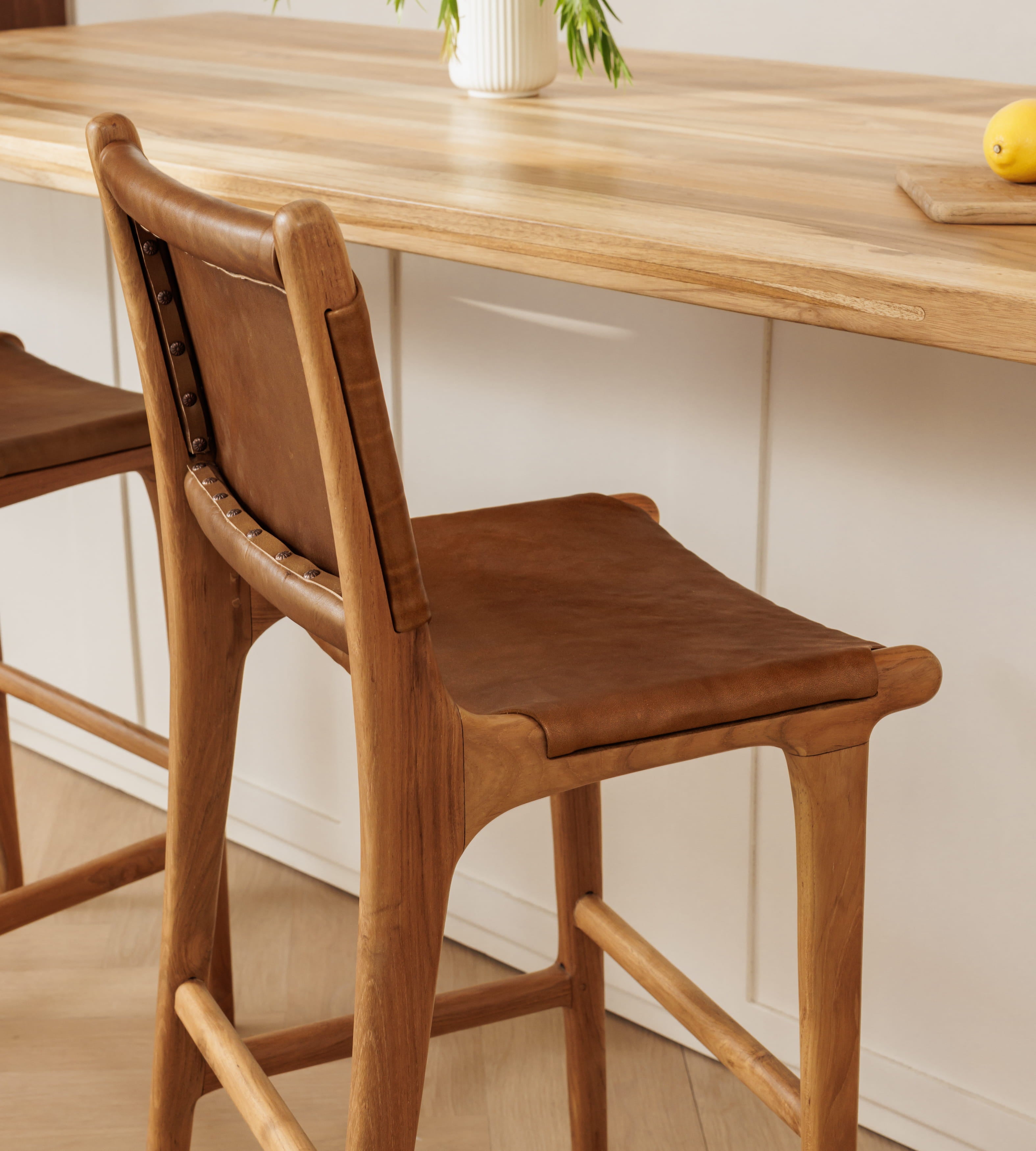 A close-up of a wooden bar stool with a brown leather seat and backrest, positioned in front of a light wood countertop in a bright, modern kitchen setting.