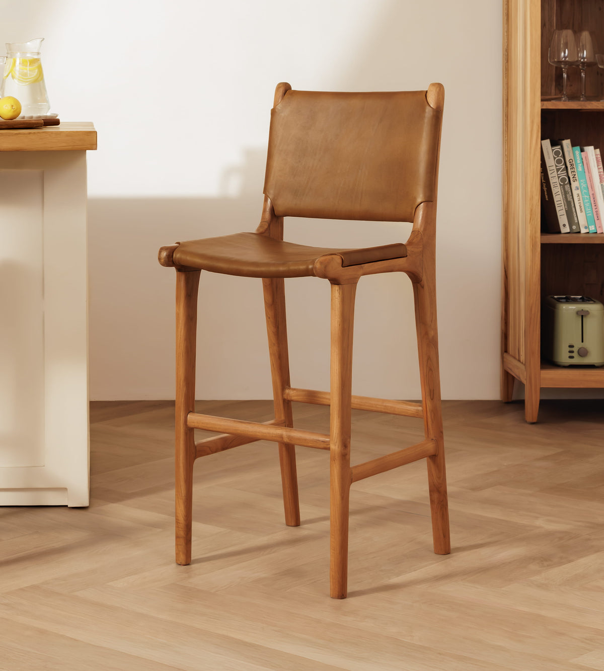A leather bar stool with a tan leather seat and backrest, shown in a warm, minimalist room setting with a wooden floor and bookshelf.