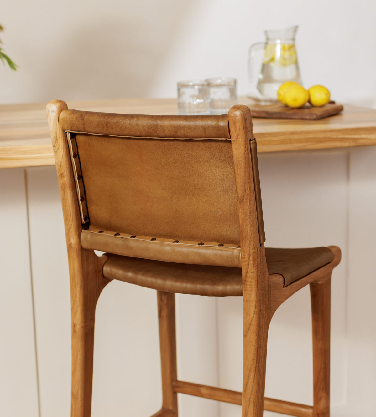 A wooden bar stool with a tan leather sling backrest and seat, shown from the back in a bright kitchen setting with a wooden countertop and glassware in the background.