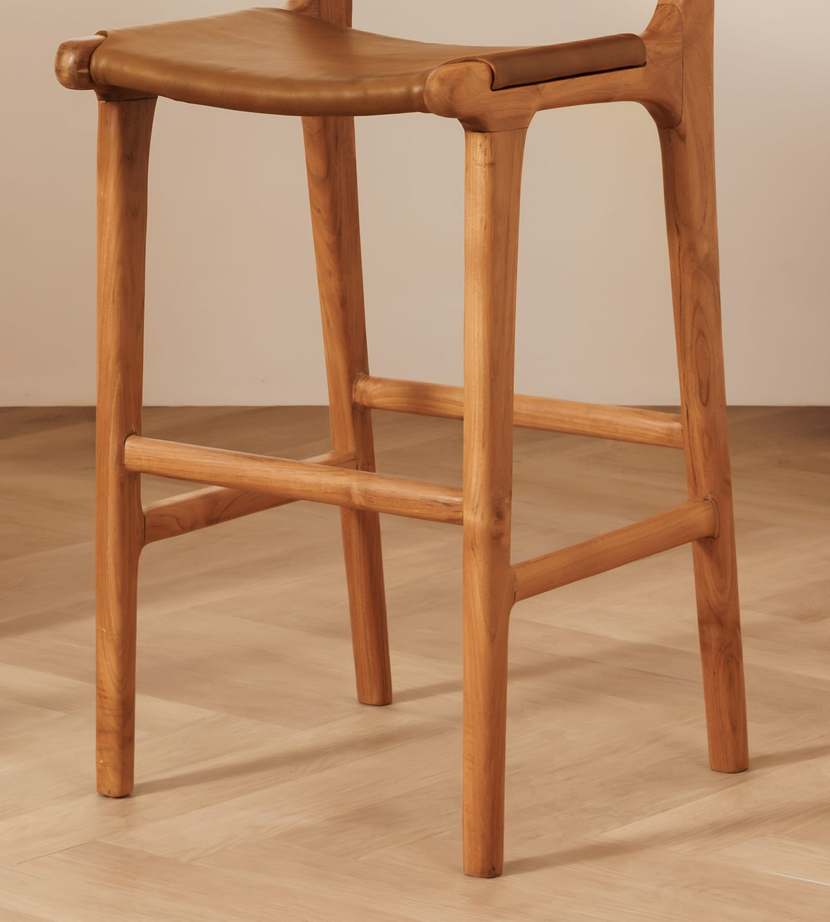 A close-up of a wooden stool with a curved backrest, woven rope seat, and natural wood legs on a light wood floor against a neutral beige wall.
