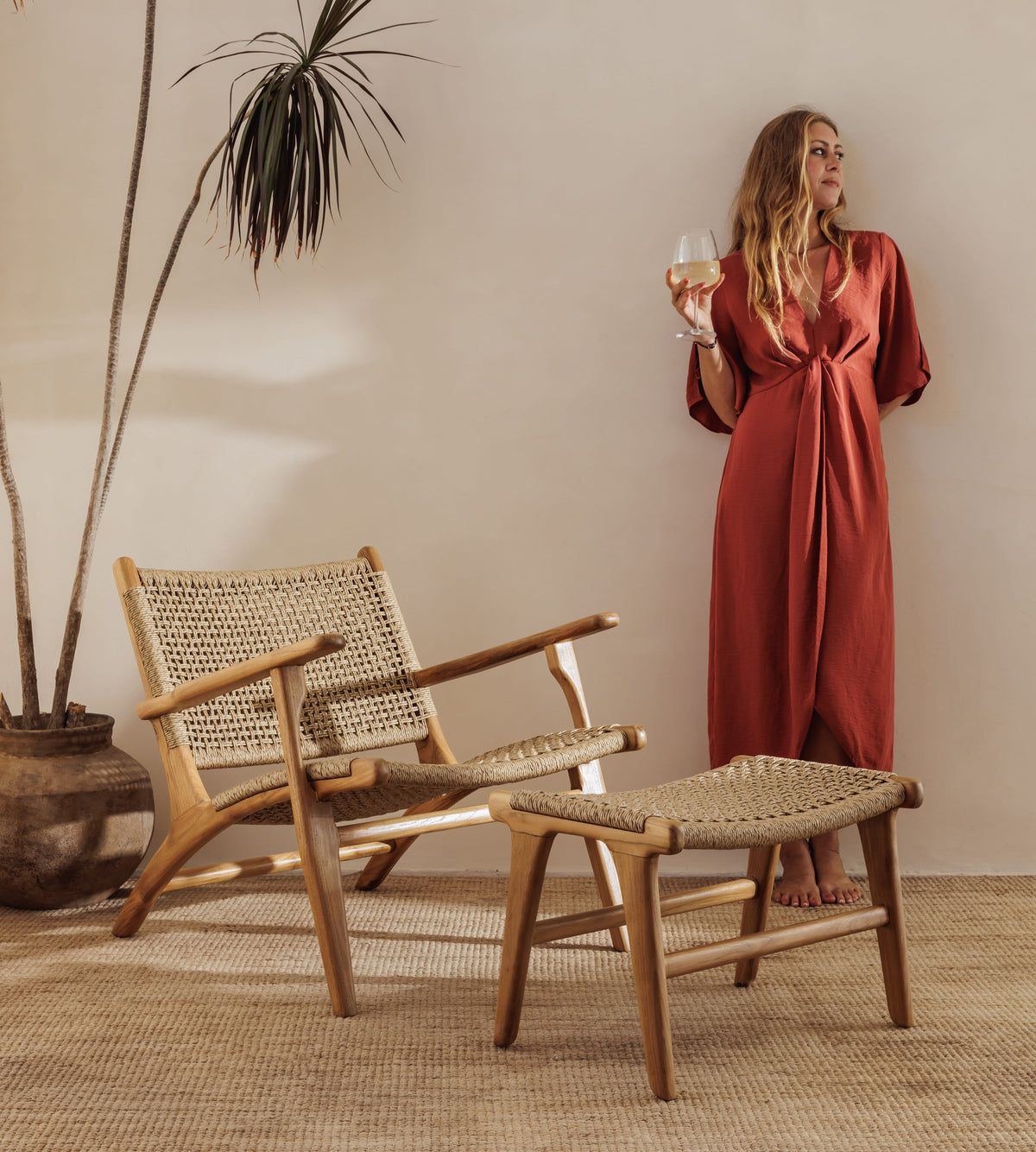 A woman in a rust-red wrap maxi dress stands barefoot in a warm, minimalist room, holding a wine glass. In the foreground are a wooden chair with a woven cane seat and a matching wooden ottoman on a jute rug.