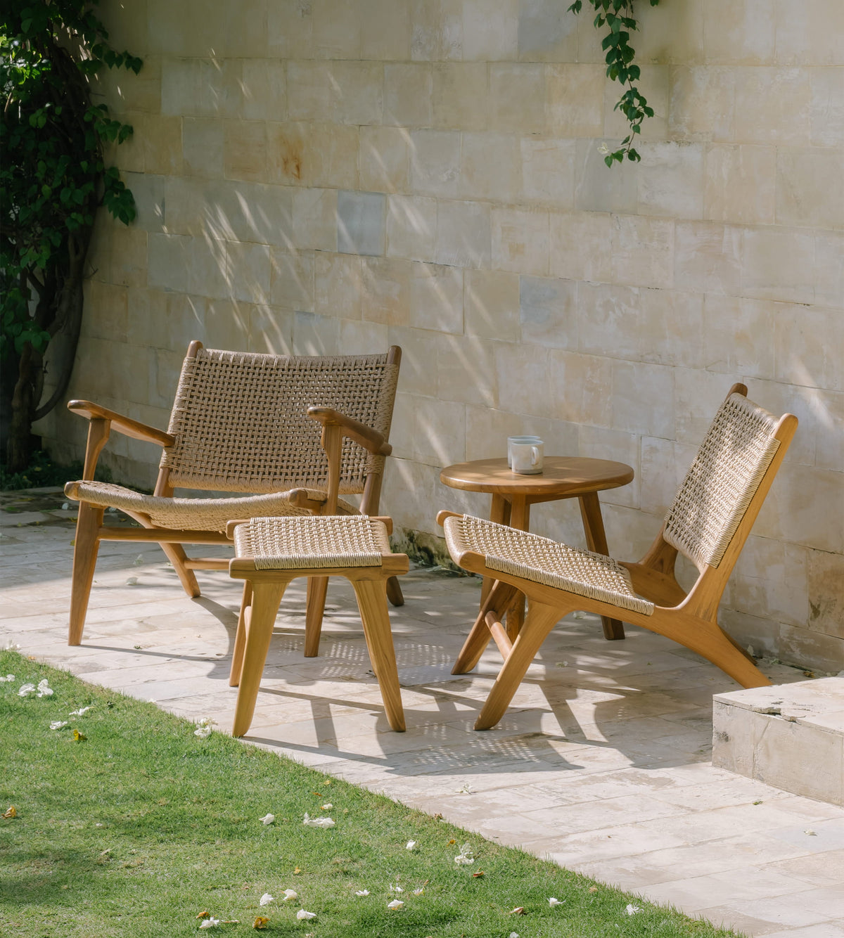 Two rattan accent chairs outdoors with woven cane seats and back, shown alongside a matching ottoman and a small round wooden side table on a stone patio against a light stone wall.