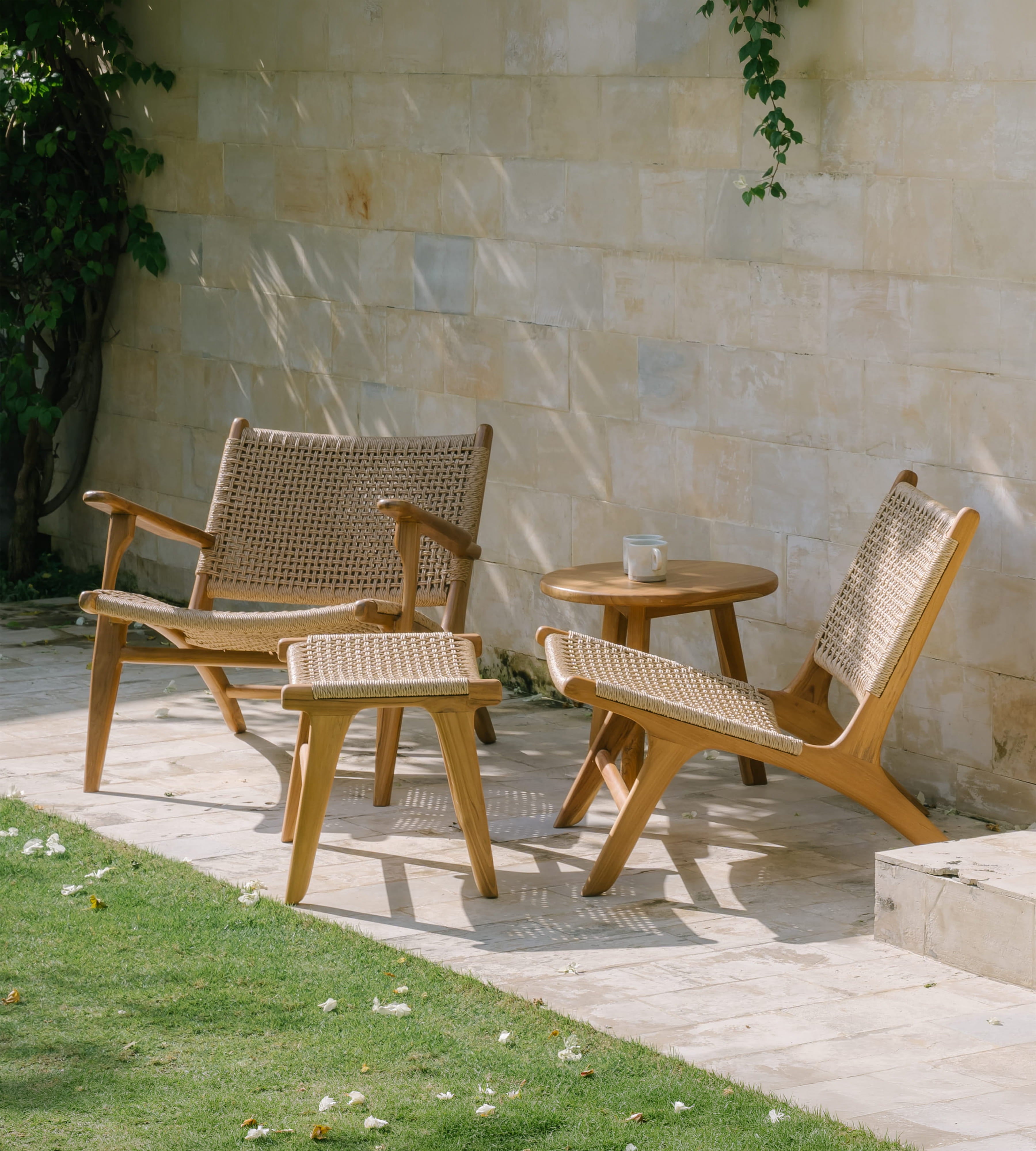 Two rattan accent chairs outdoors with woven cane seats and back, shown alongside a matching ottoman and a small round wooden side table on a stone patio against a light stone wall.