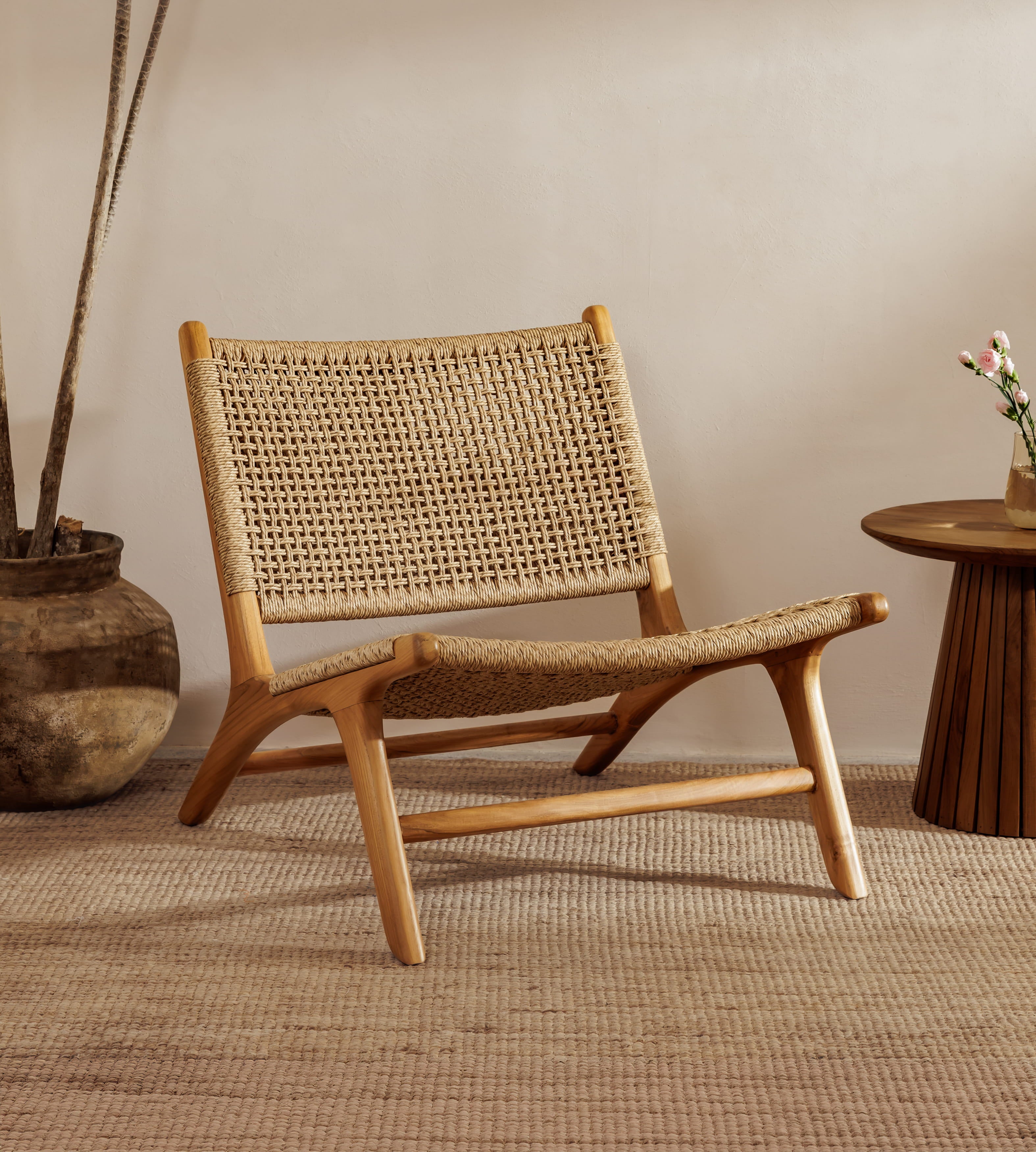 A rattan accent chair chair with a natural wood frame and woven rattan backrest, styled in a warm, minimalist room with a woven rug, a potted plant, and a small wooden side table.