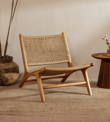 A rattan accent chair chair with a natural wood frame and woven rattan backrest, styled in a warm, minimalist room with a woven rug, a potted plant, and a small wooden side table.