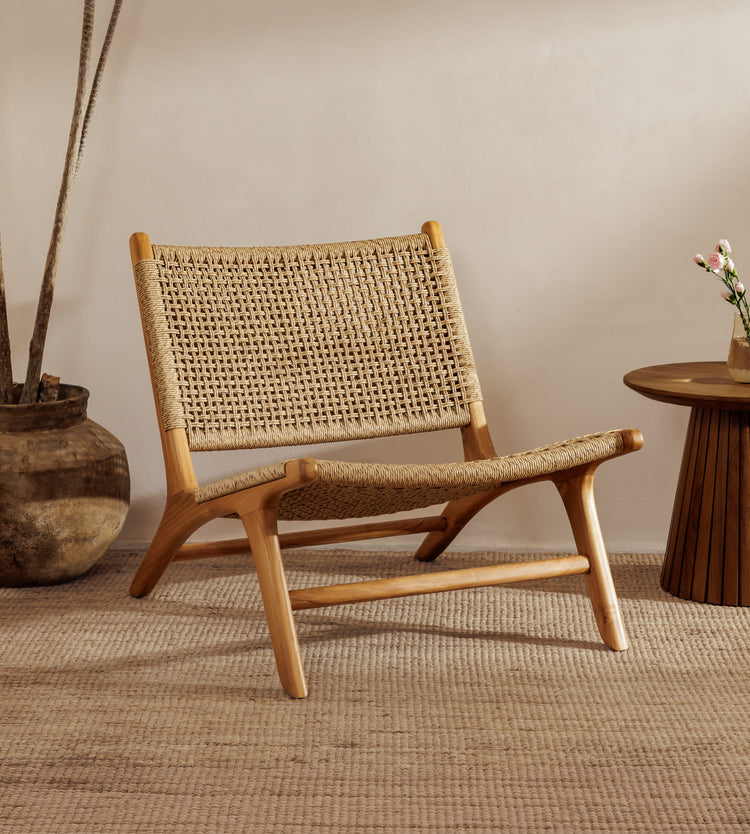 A rattan accent chair chair with a natural wood frame and woven rattan backrest, styled in a warm, minimalist room with a woven rug, a potted plant, and a small wooden side table.