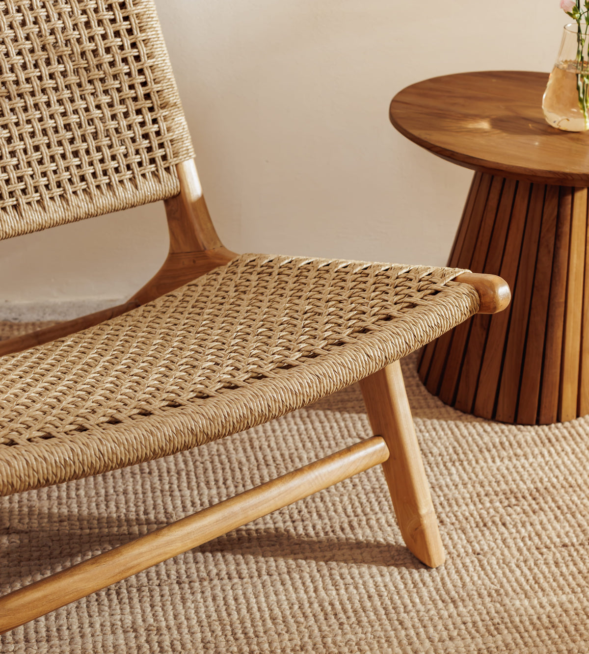A close-up of a mid-century modern wooden lounge chair with woven natural fiber seat and back, shown in a warm, neutral-toned interior next to a round wooden side table.