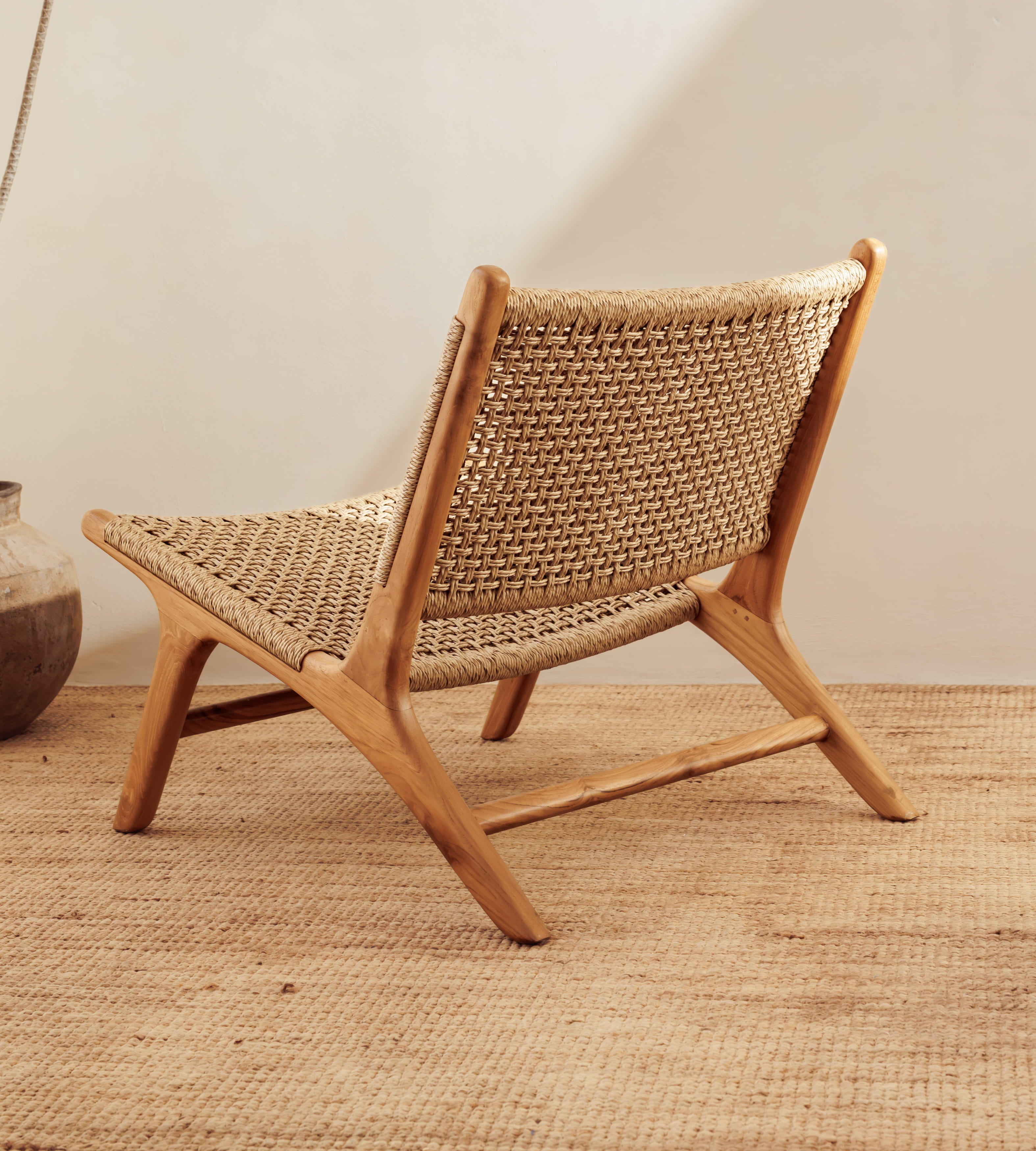 A mid-century modern wooden lounge chair with a woven cane or rattan seat and back, shown in a styled interior setting on a textured jute rug.