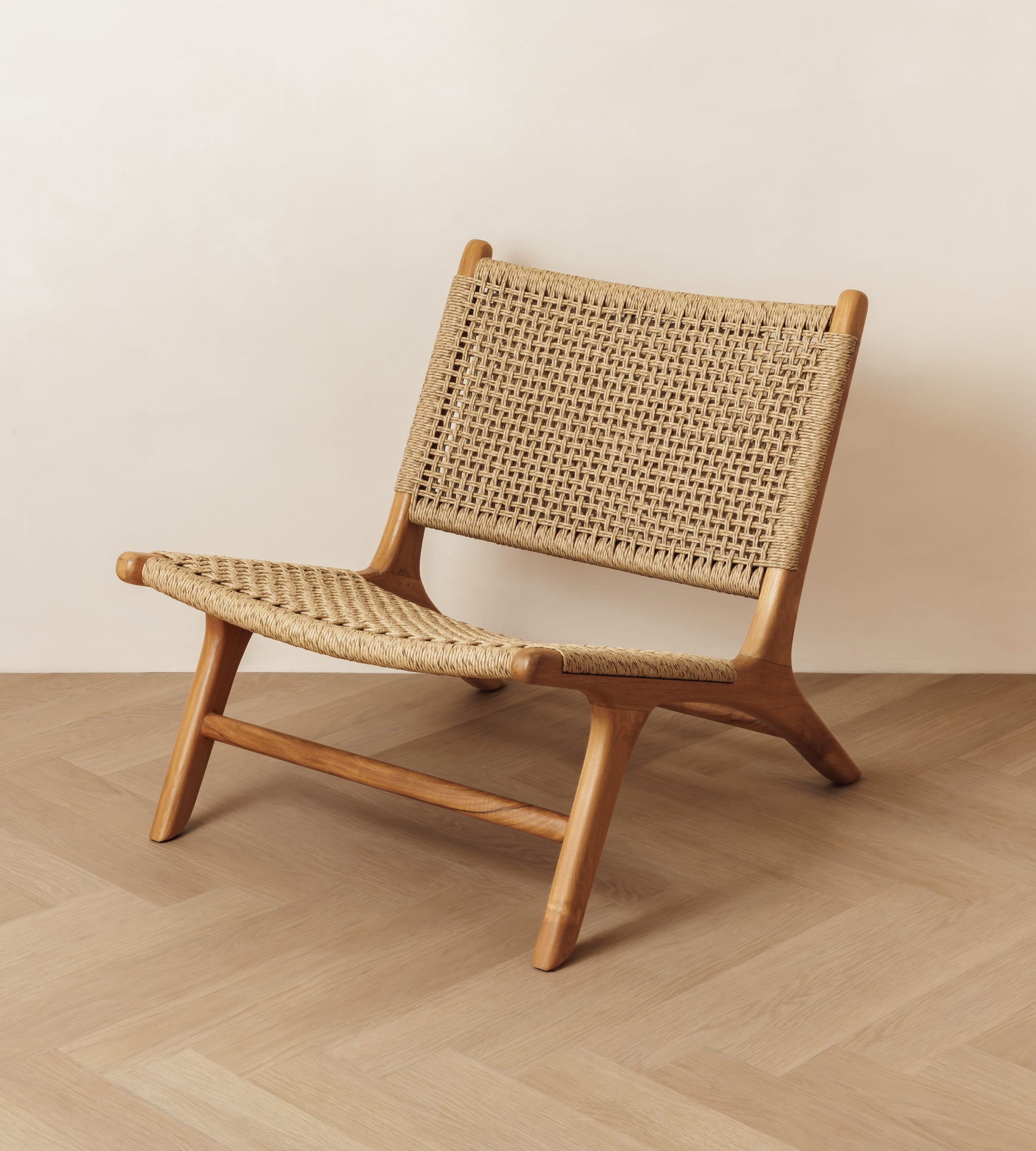 A mid-century modern rattan lounge chair with woven cane or rattan seat and back, shown on a light wood herringbone floor against a plain beige wall.