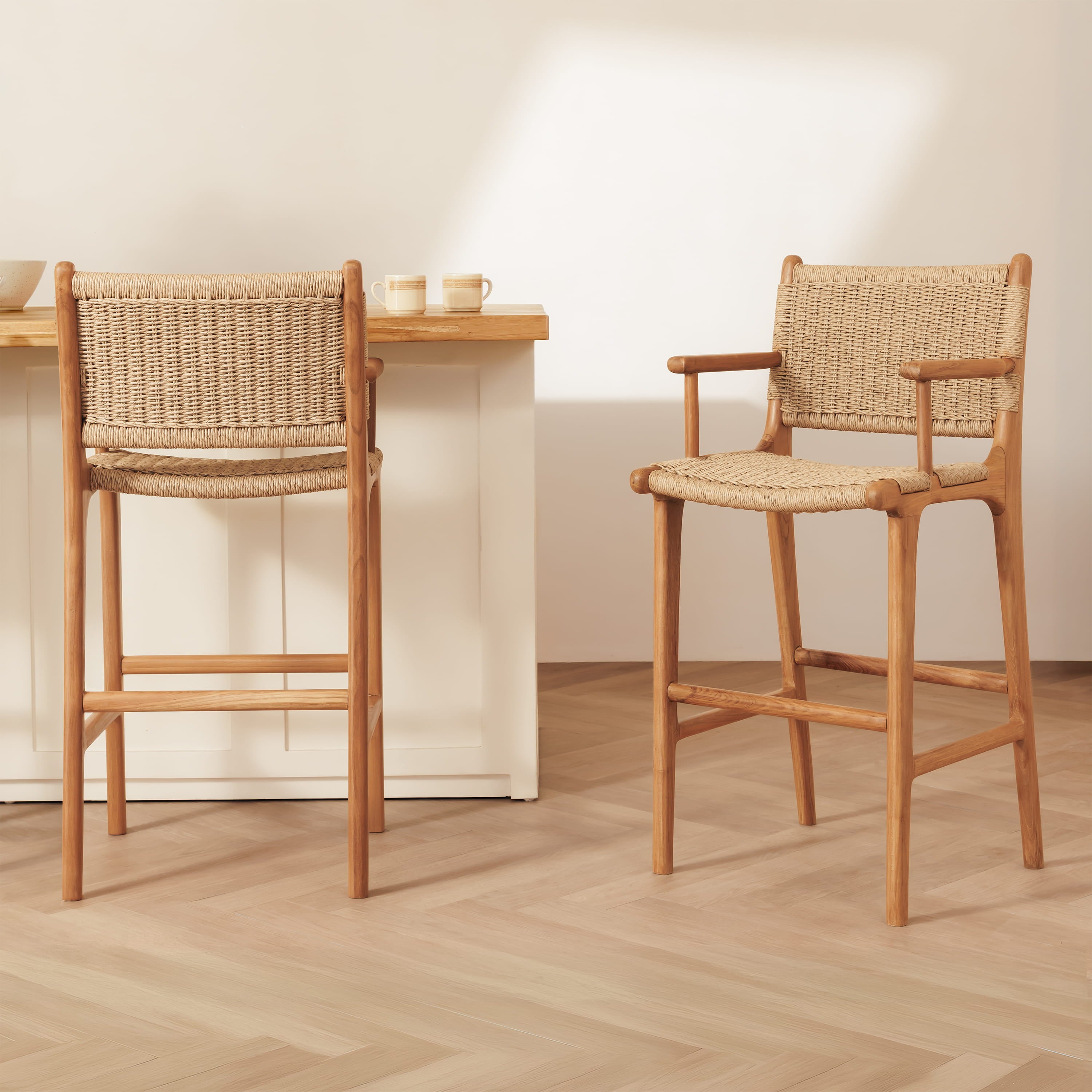 Two wooden bar stools with woven rope seats and backs, shown in a bright minimalist interior with a light wood floor and white wall.