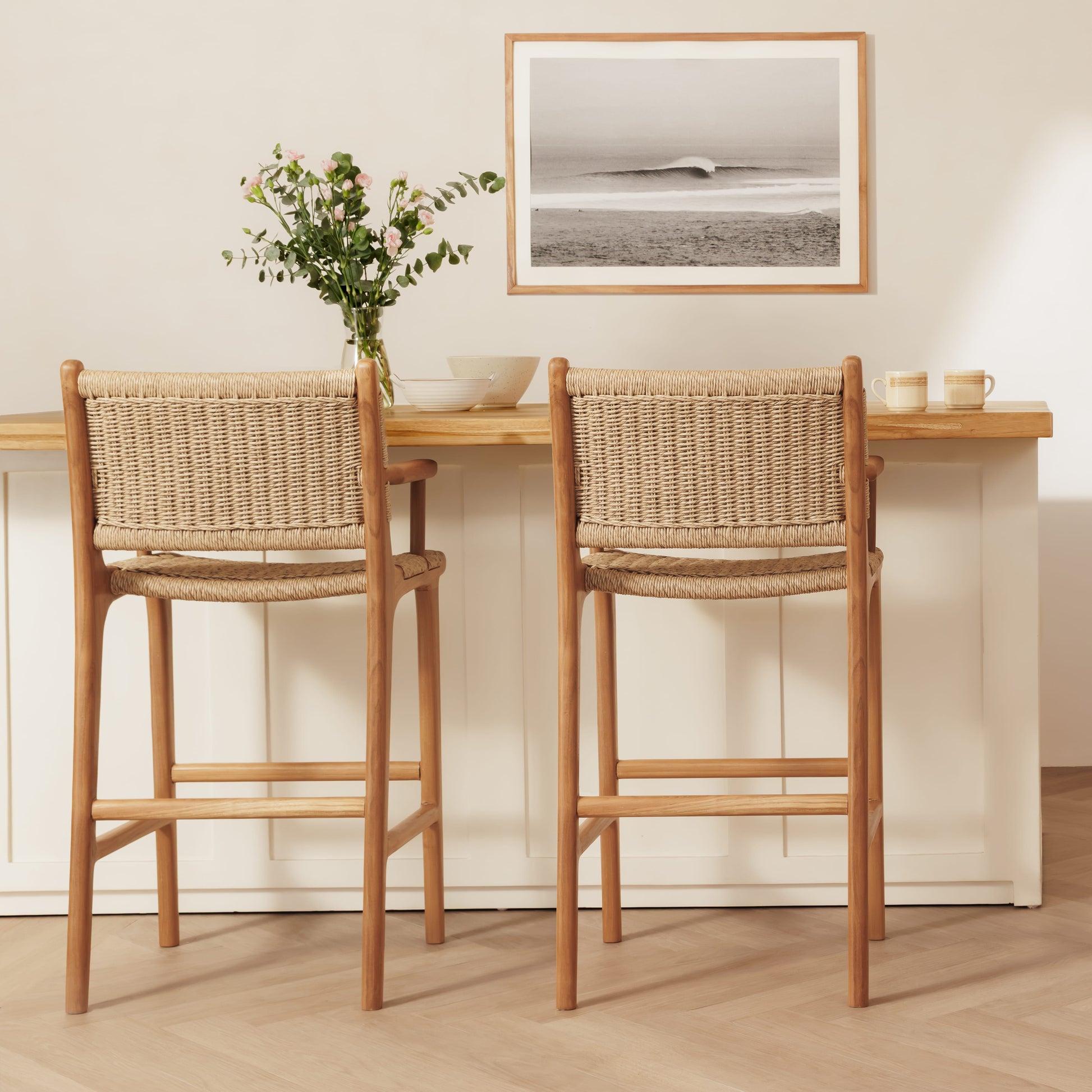 Two wooden bar stools with woven rope backs and seats, shown in a bright, modern kitchen setting with a light wood counter and a framed print on the wall.