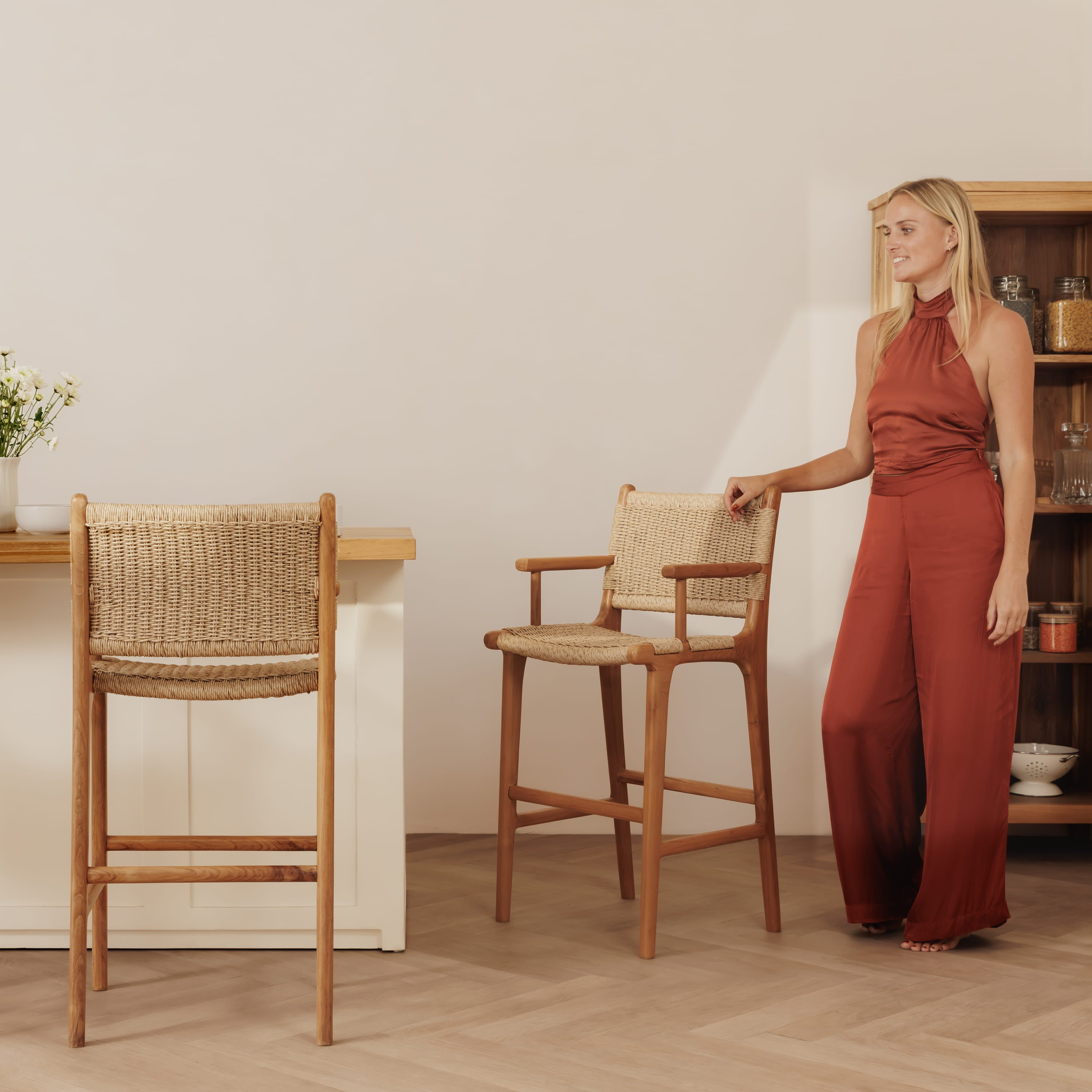 A woman in a rust-colored halter jumpsuit stands beside a wooden bar stool with a woven cane seat and back in a bright, modern room. Another matching stool is visible on the left.