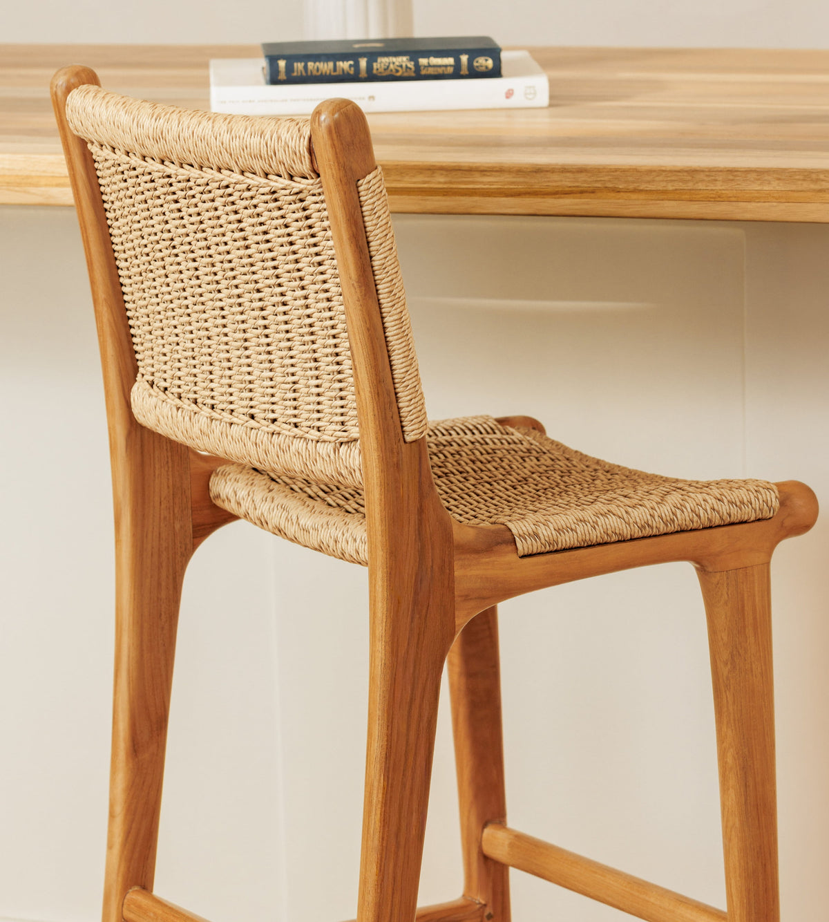 A wooden bar stool with a woven rattan seat and back, shown in a warm indoor setting with a wooden table and books in the background.