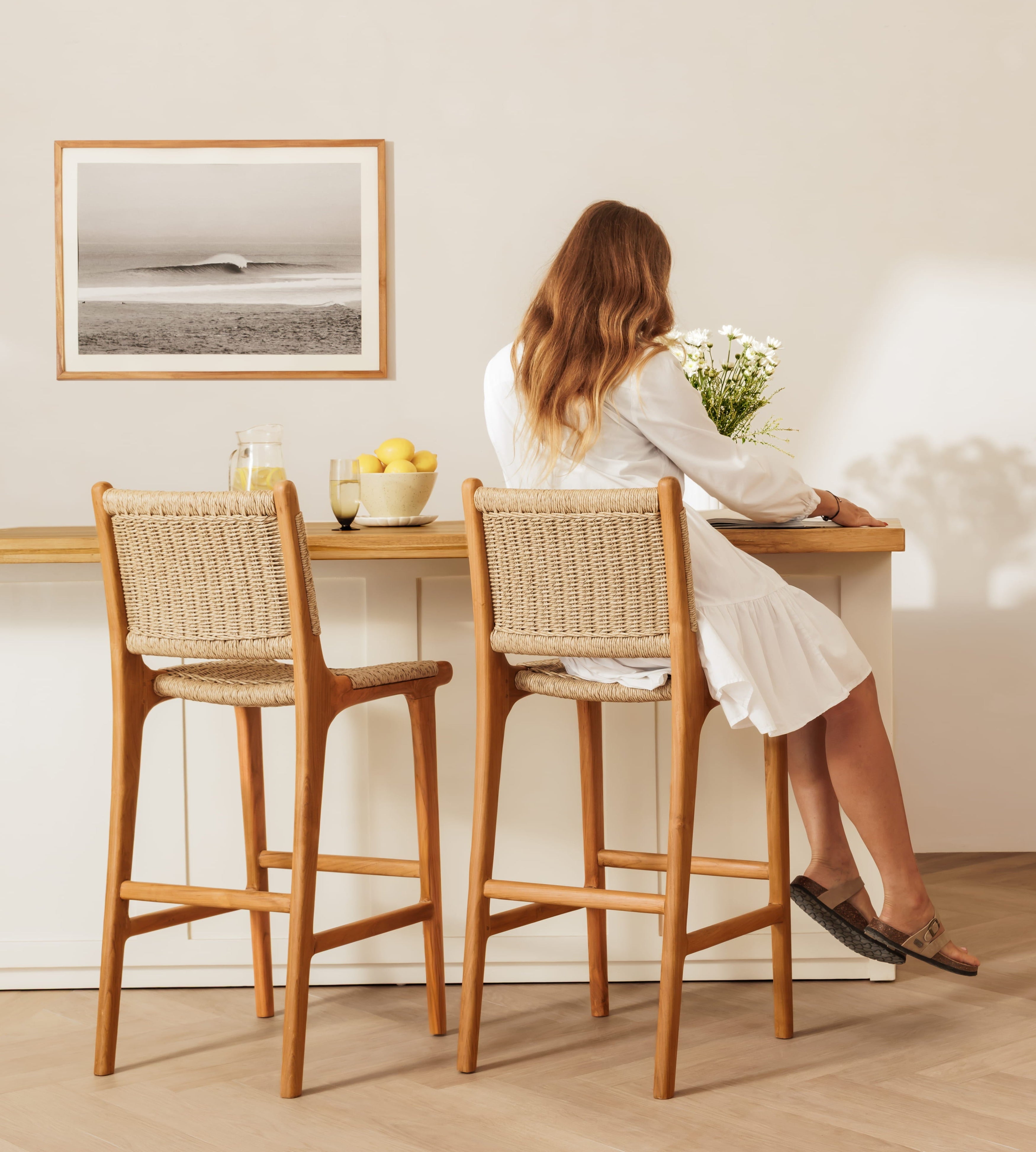 A woman sits at a wooden bar-height rattan bar stool in a bright, modern room, viewed from behind. The stool has a natural woven cane back and seat with a light wood frame.