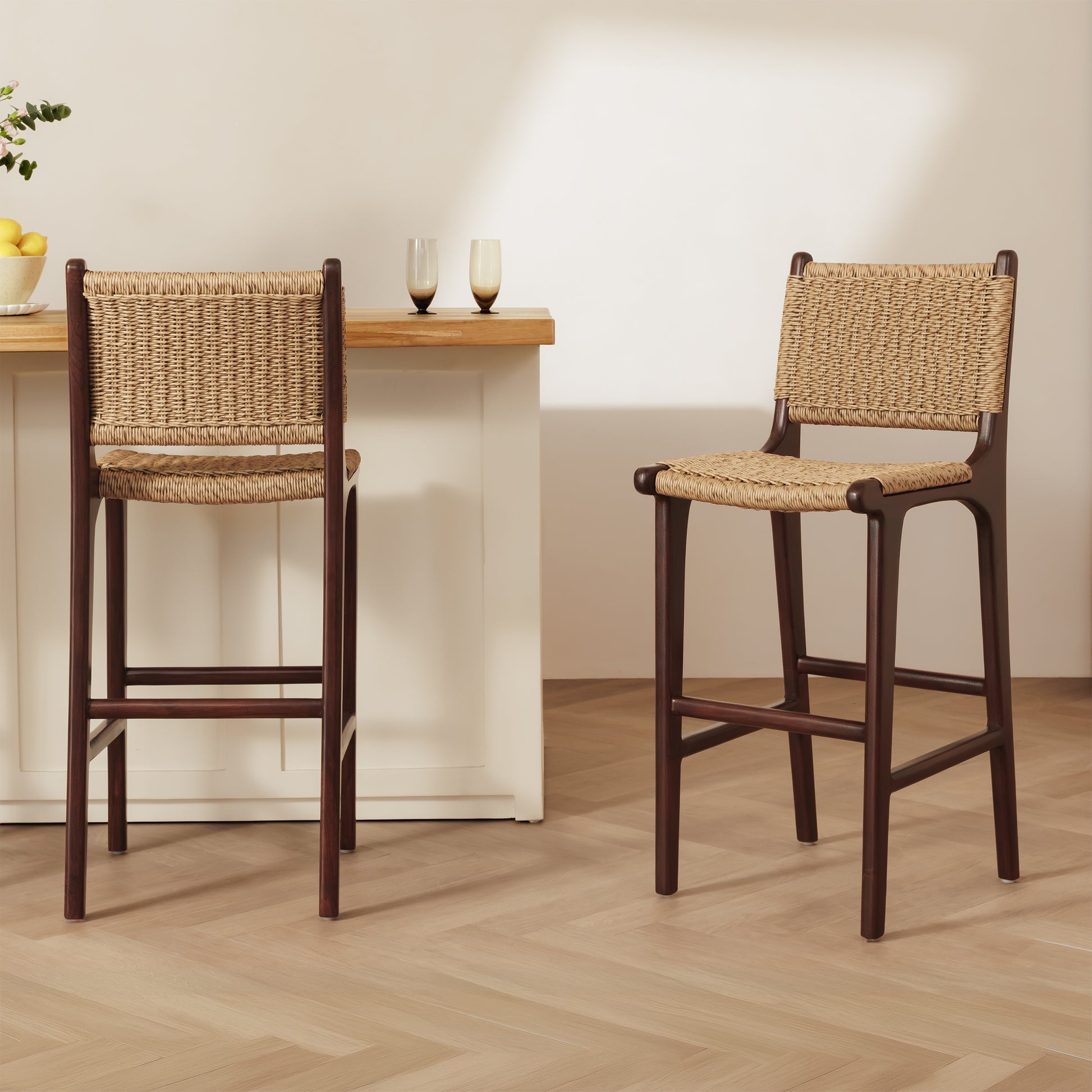 Two dark rattan bar stools with woven rattan seats and backs, shown in a bright kitchen or dining setting with a light wood counter and pale wall.