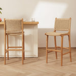 Two rattan bar stools with woven rattan seats and backs, shown in a bright, modern interior with a light wood table and pale wall.