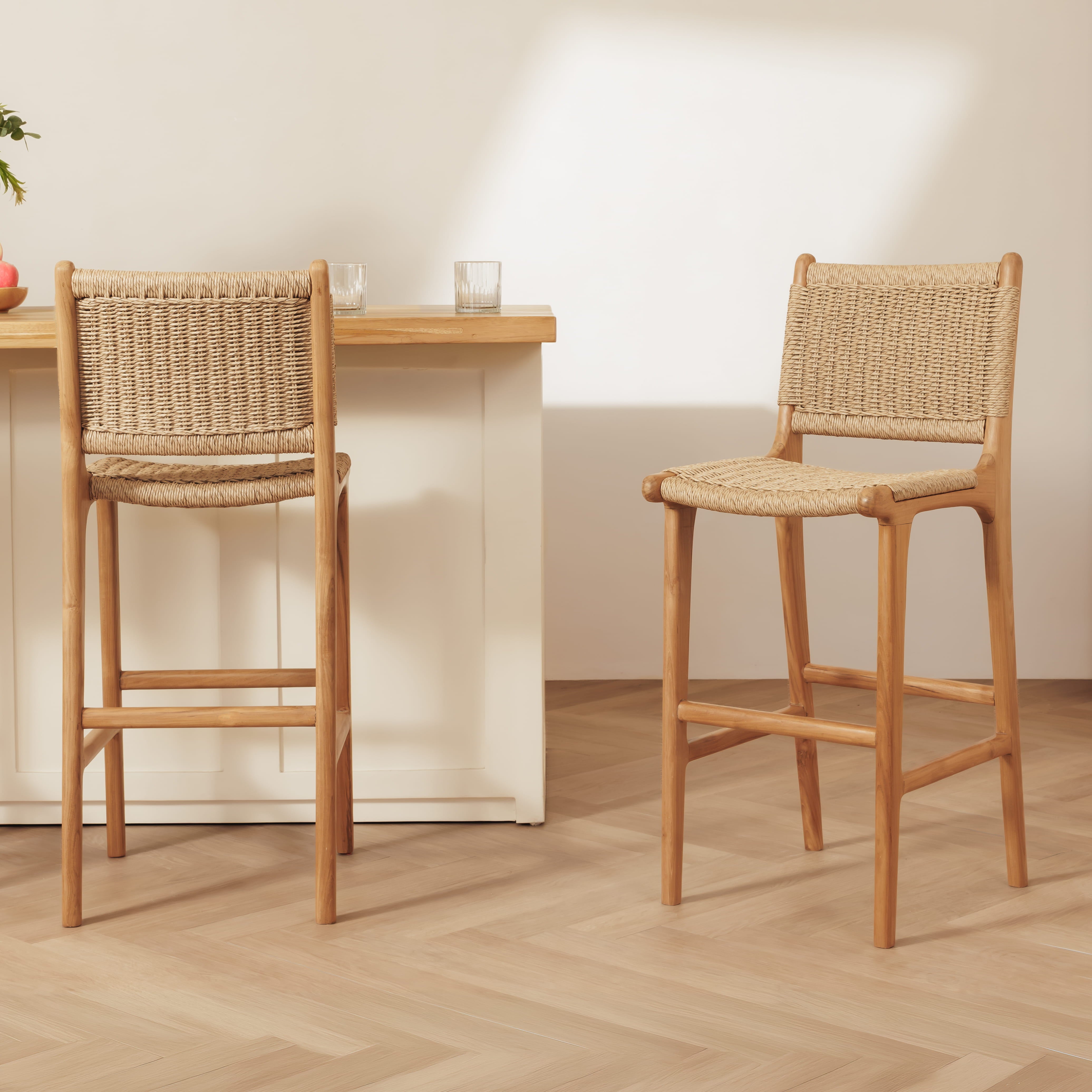 Two rattan bar stools with woven rattan seats and backs, shown in a bright, modern interior with a light wood table and pale wall.