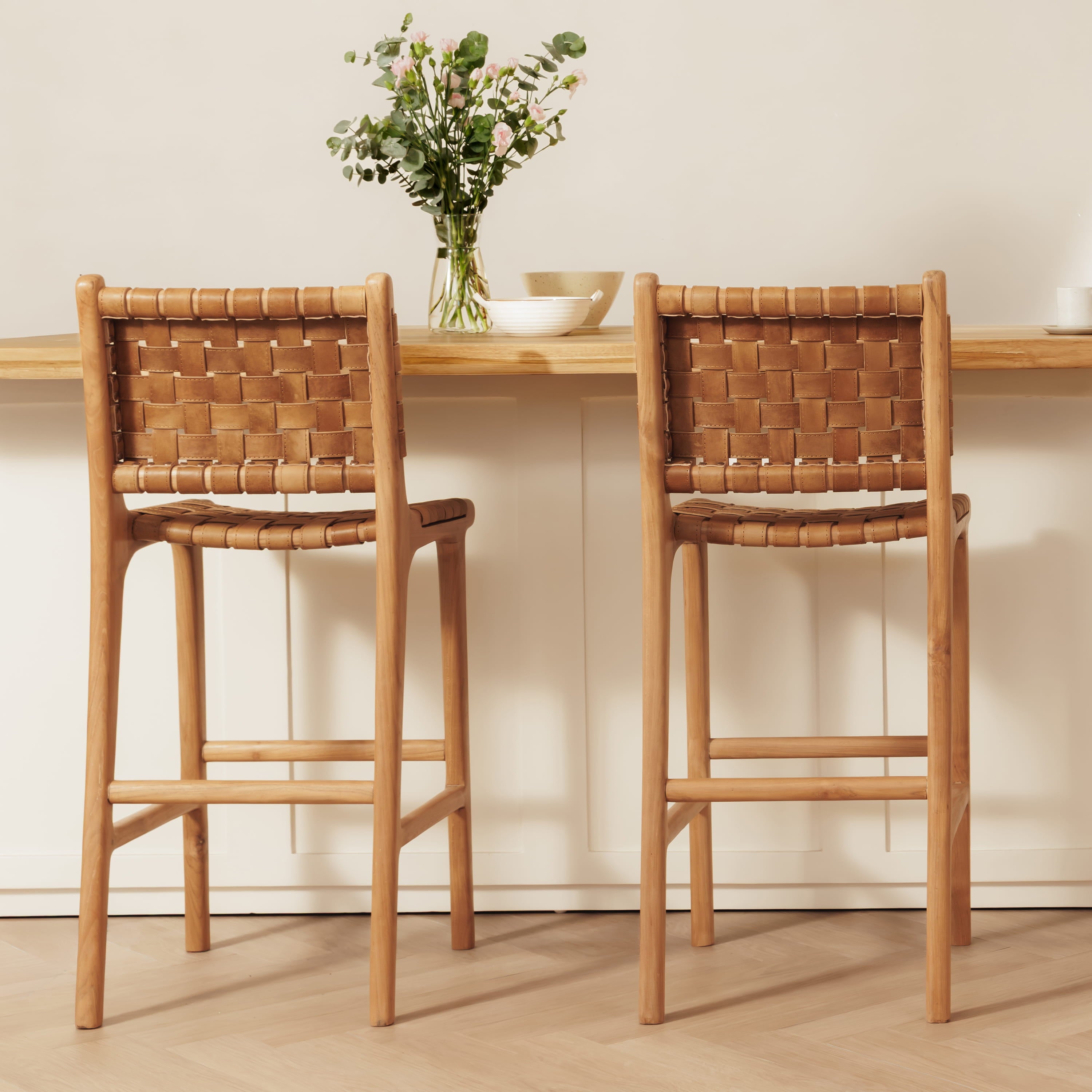 Two wooden bar stools with woven leather backs and seats, positioned in front of a light wood counter with a vase of greenery.