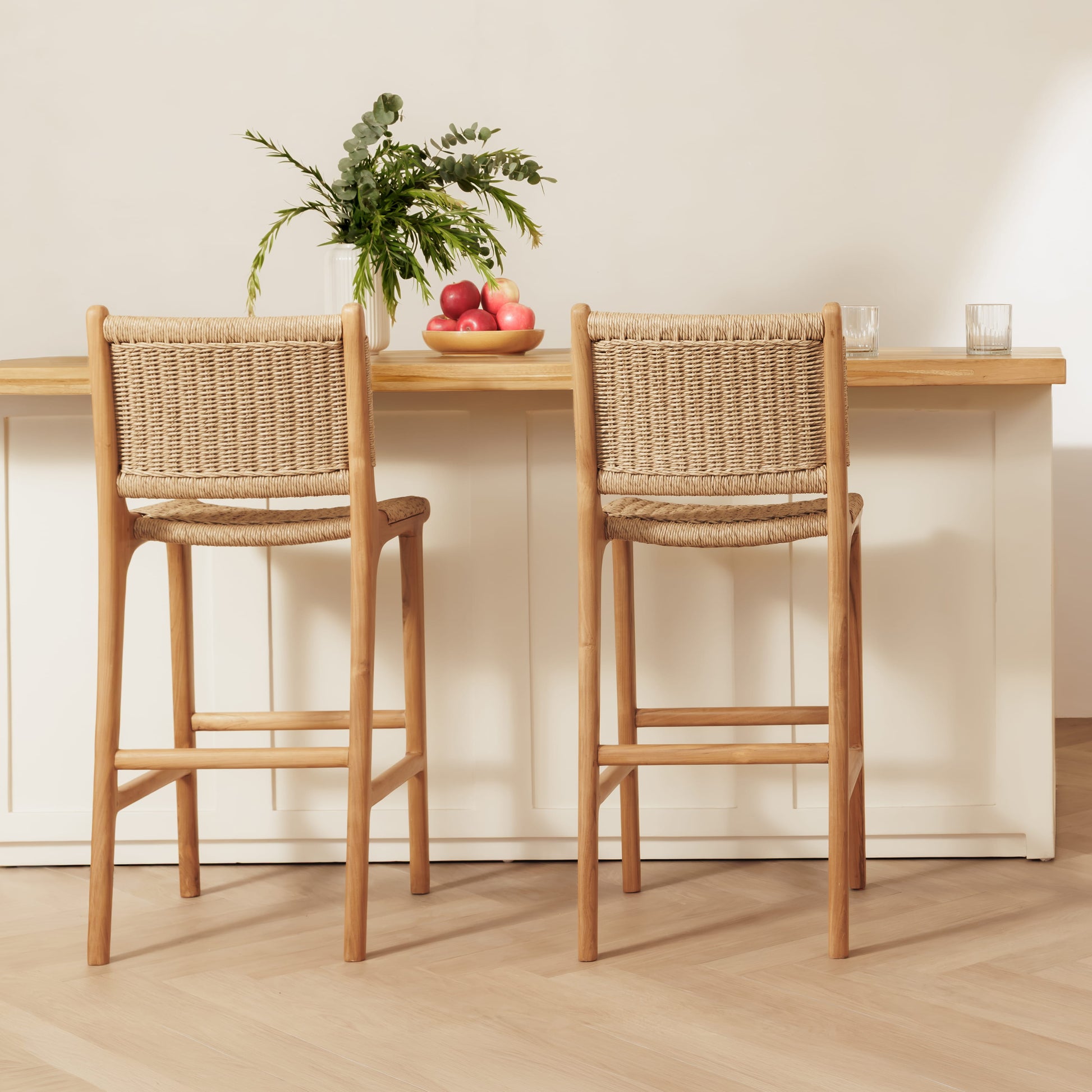 Two rattan bar stools with rattan rope seats and backs, positioned in front of a light wood and white counter with a vase of greenery and fruit on top.