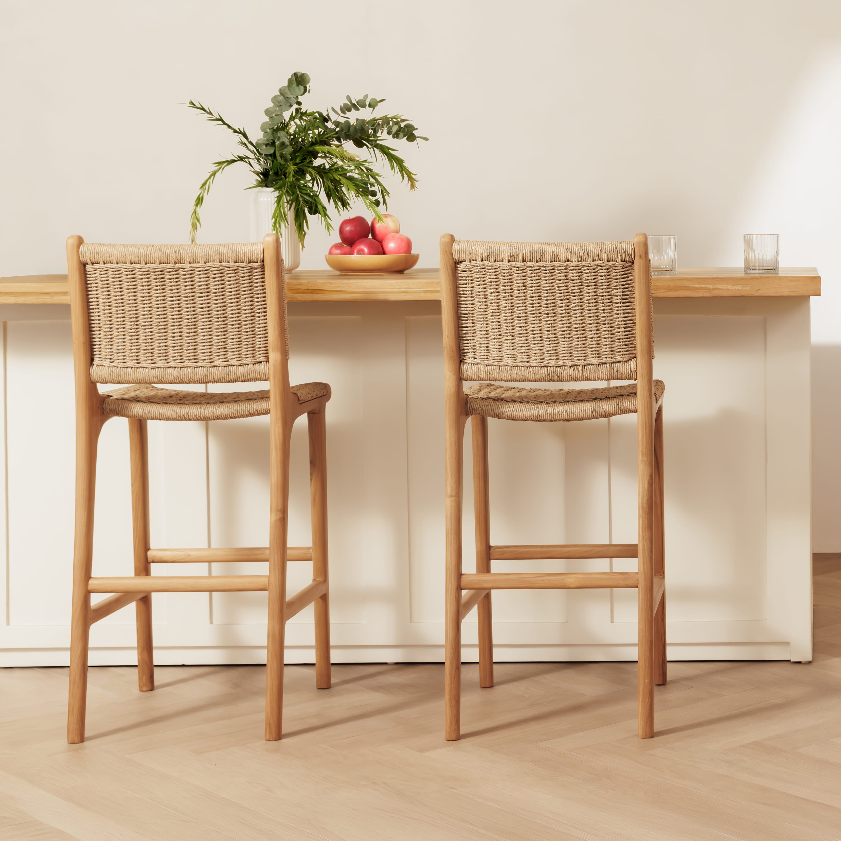 Two rattan bar stools with rattan rope seats and backs, positioned in front of a light wood and white counter with a vase of greenery and fruit on top.