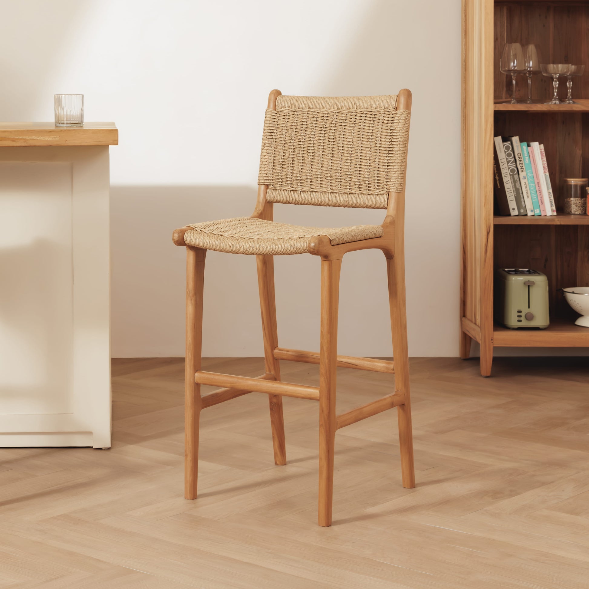 A rattan bar stool with a woven rattan seat and backrest, shown in a bright, modern room setting with a light wood floor, a white table on the left, and a wooden bookshelf on the right.