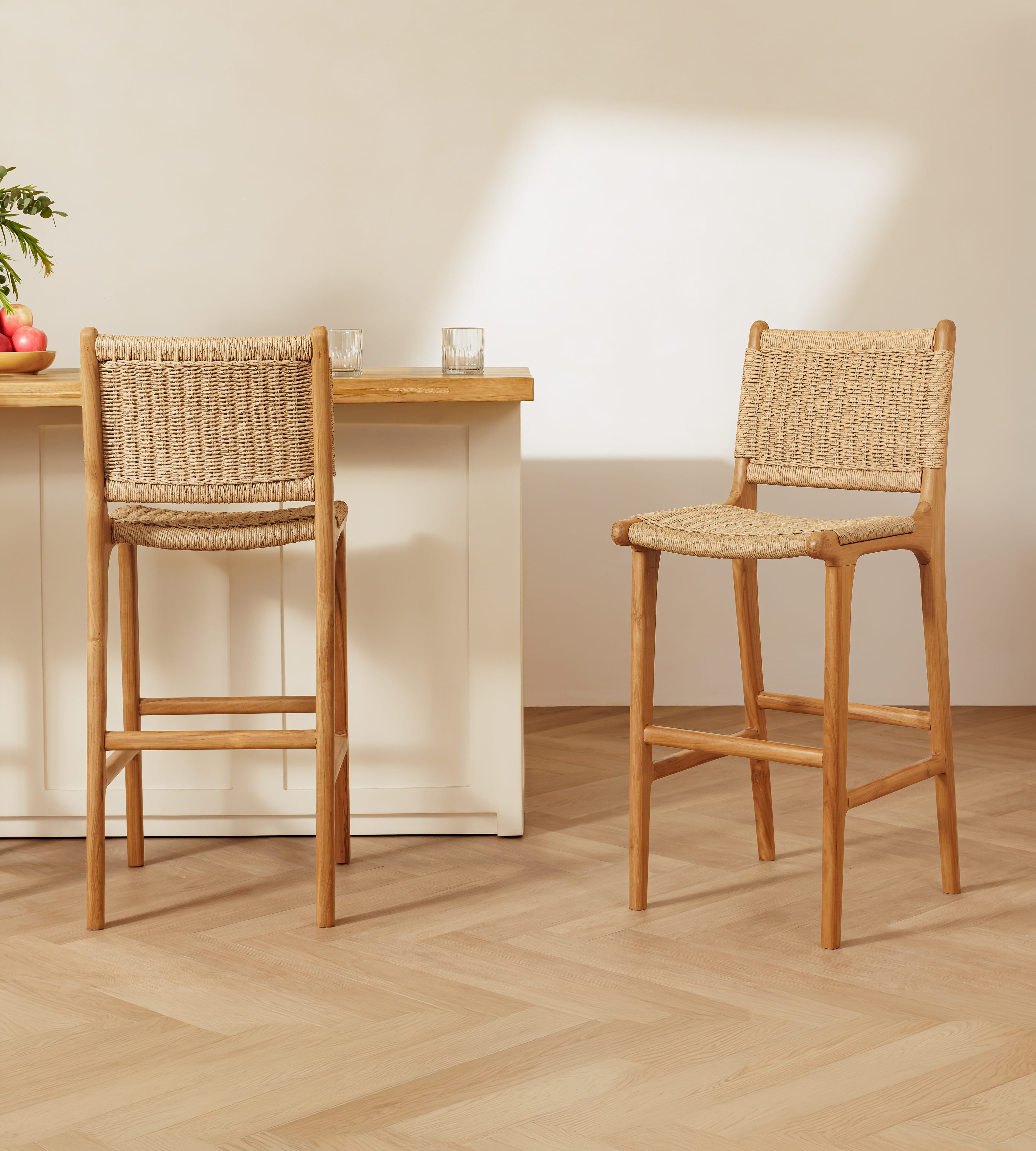 Two rattan bar stools with woven rattan seats and backs, shown in a bright, modern kitchen setting with a light wood table and white cabinets.