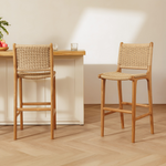 Two rattan bar stools with woven rattan seats and backs, shown in a bright, modern kitchen setting with a light wood table and white cabinets.