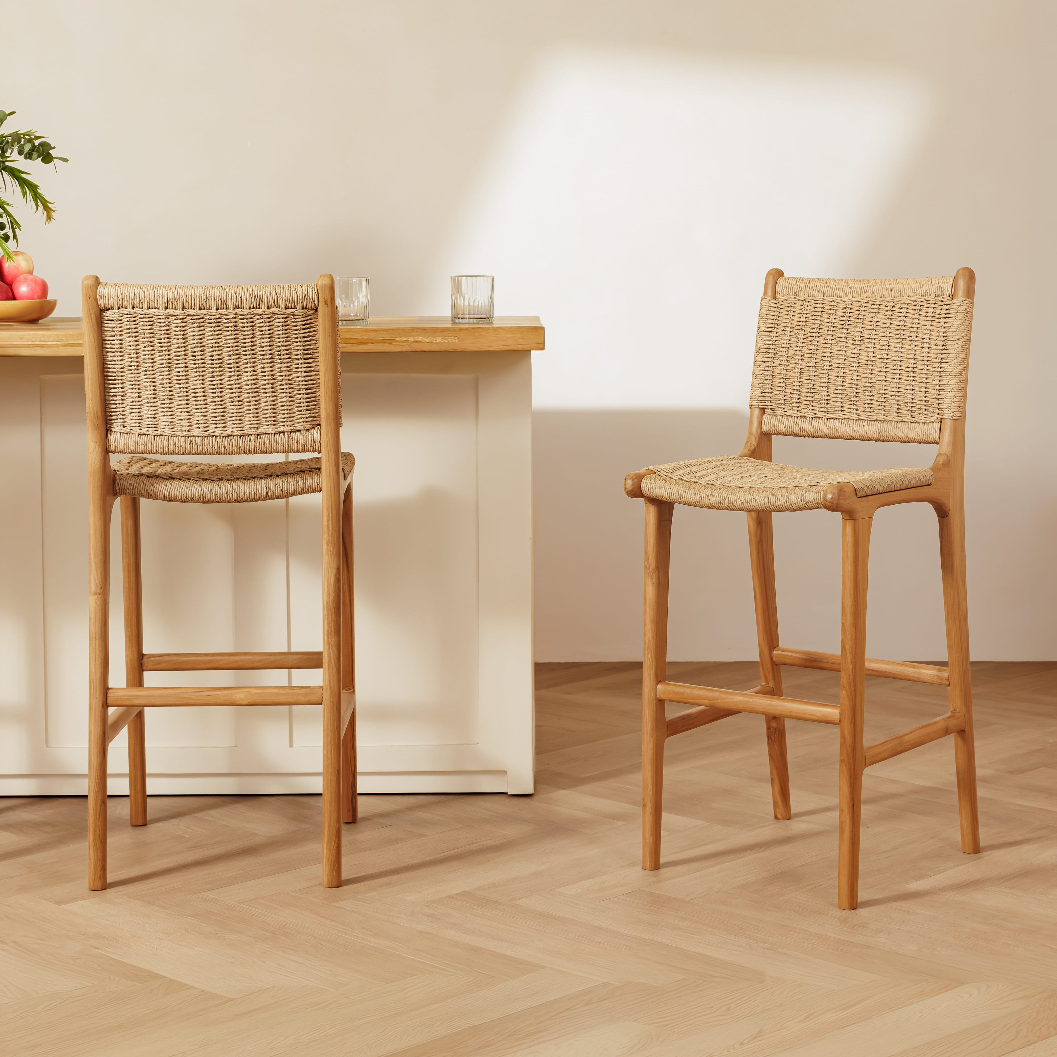 Two rattan bar stools with woven rattan seats and backs, shown in a bright, modern kitchen setting with a light wood table and white cabinets.