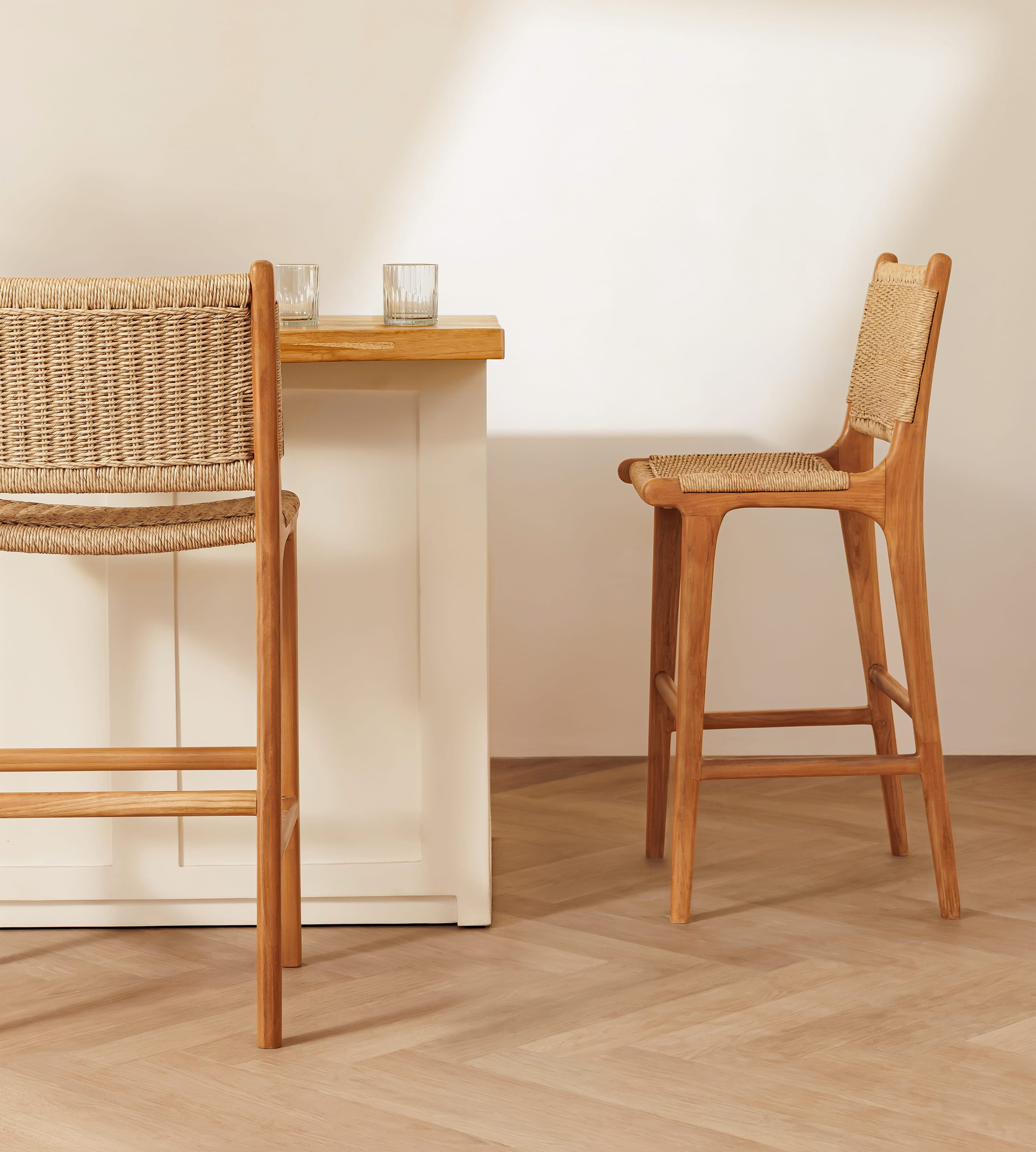 Two wooden bar stools with woven rope seats and backs, shown in a bright minimalist interior with a white table and light wood floor.