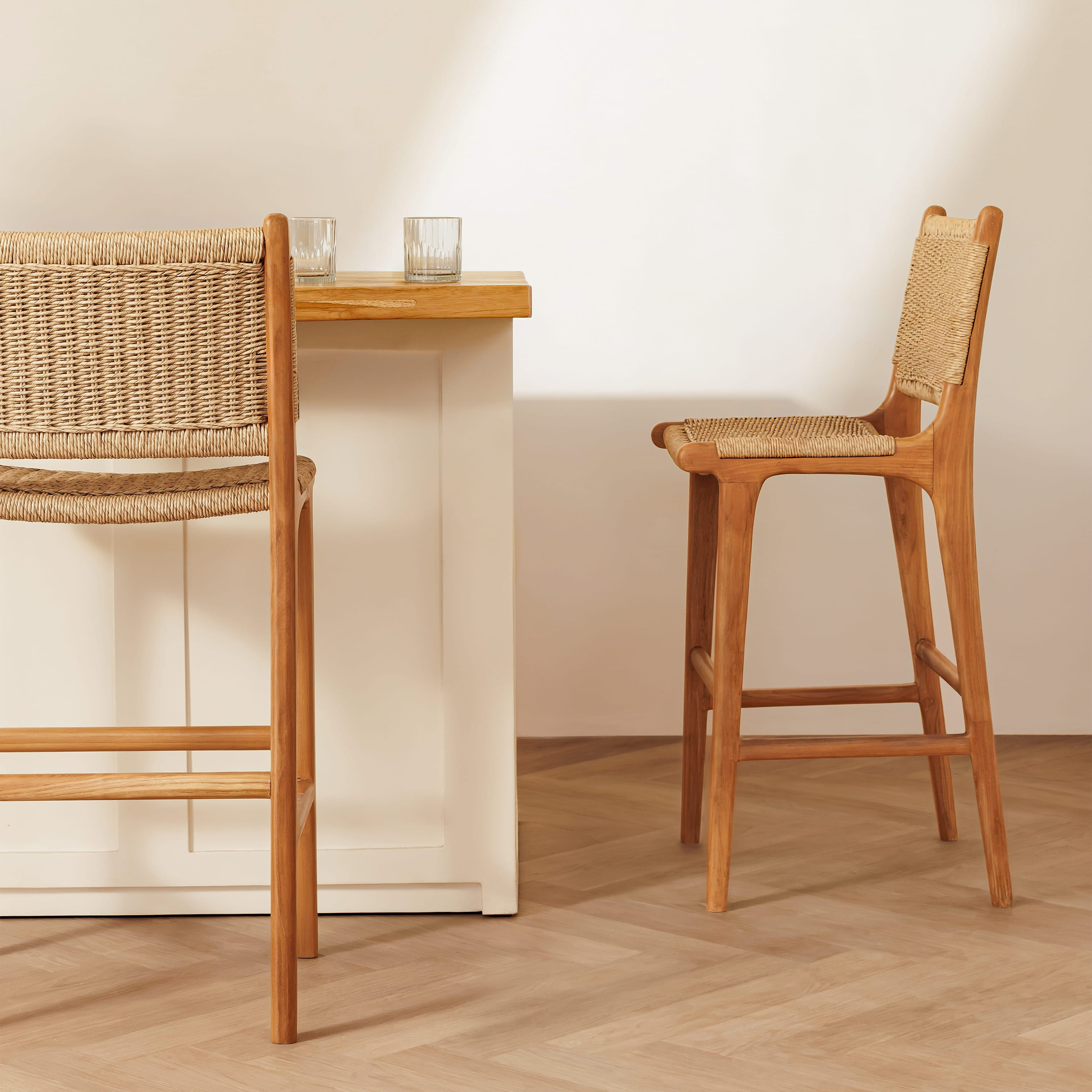 Two wooden bar stools with woven rope seats and backs, shown in a bright minimalist interior with a white table and light wood floor.