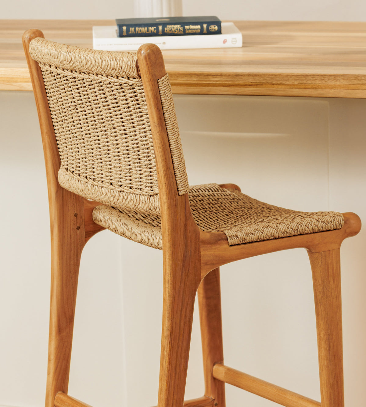 A wooden bar stool with a woven rattan seat and back, shown in a warm indoor setting with a wooden table and books in the background.