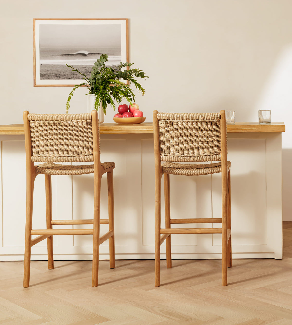 Two rattan bar stools with woven rattan seats and backs, positioned in front of a light wood and white counter in a bright, modern interior.