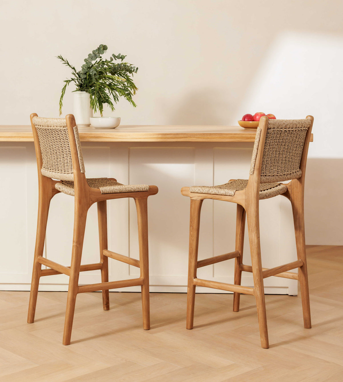 Two rattan bar stools with woven cane backs and seats, positioned in front of a light wood counter in a bright, modern interior.