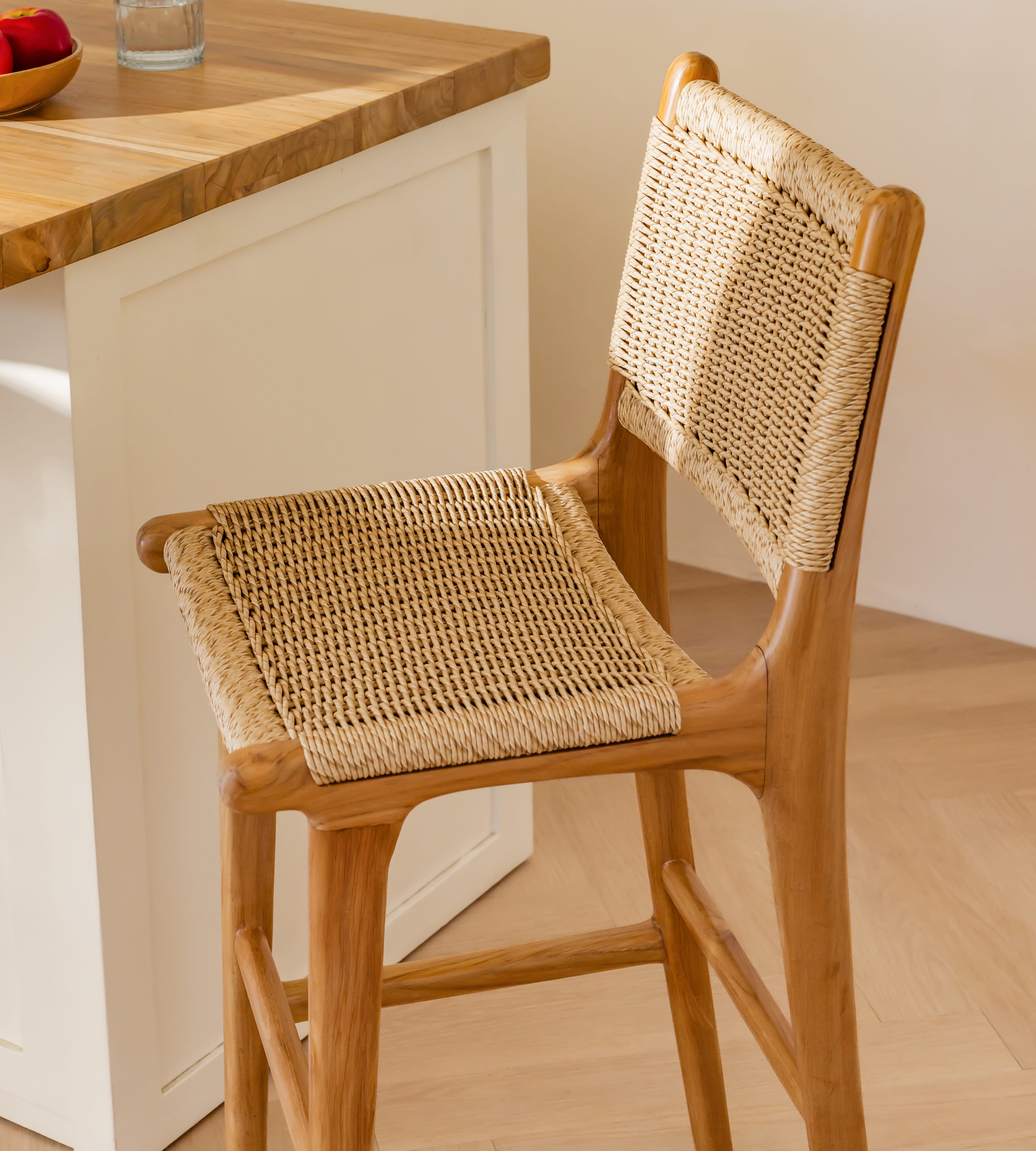 A wooden bar stool with a woven rattan seat and back, shown in a bright kitchen setting next to a wooden countertop.