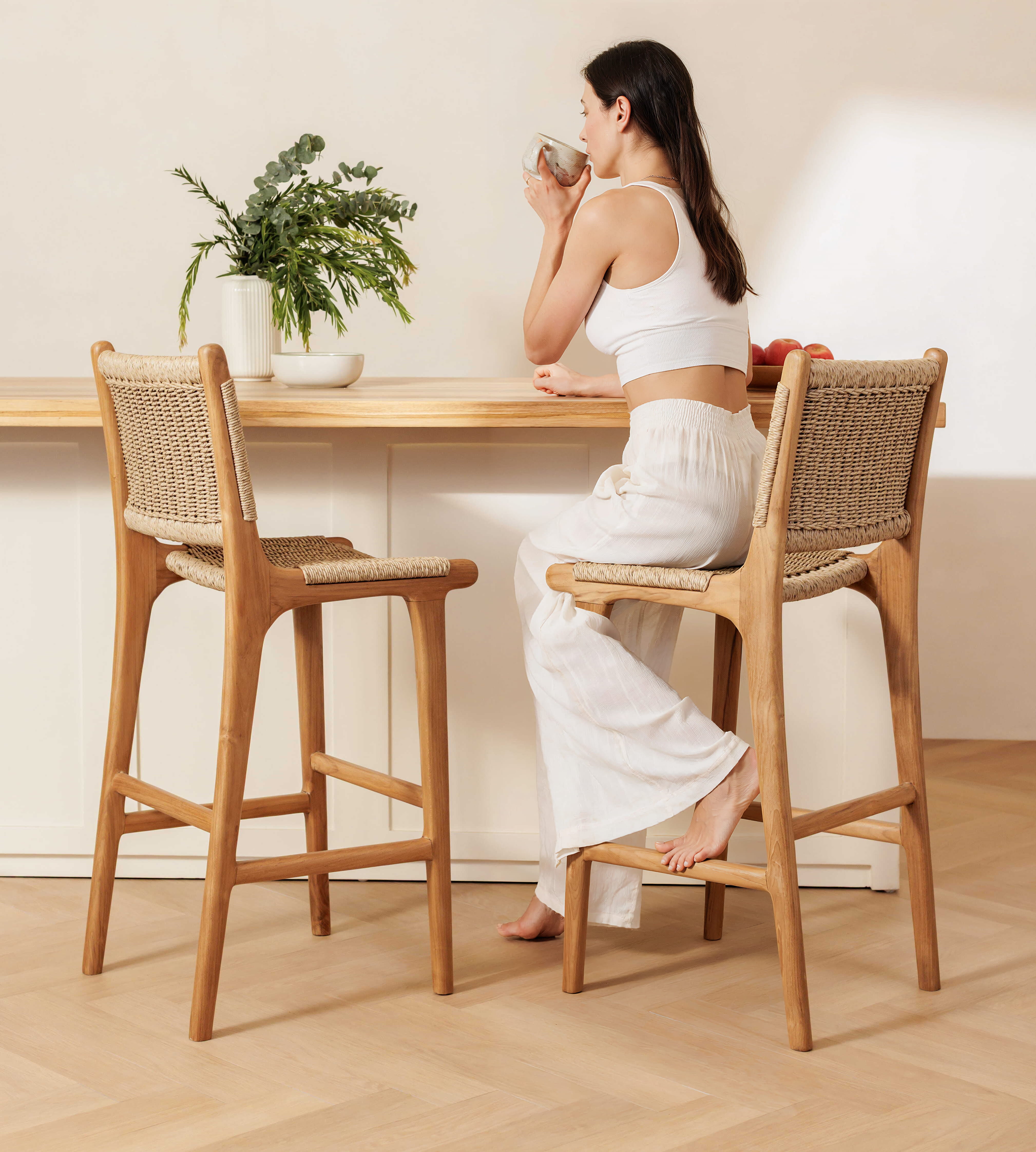 A woman sits barefoot on a wooden bar stool with a woven seat and back, positioned beside a matching wooden counter in a bright, minimalist interior. Another matching stool stands empty on the left.