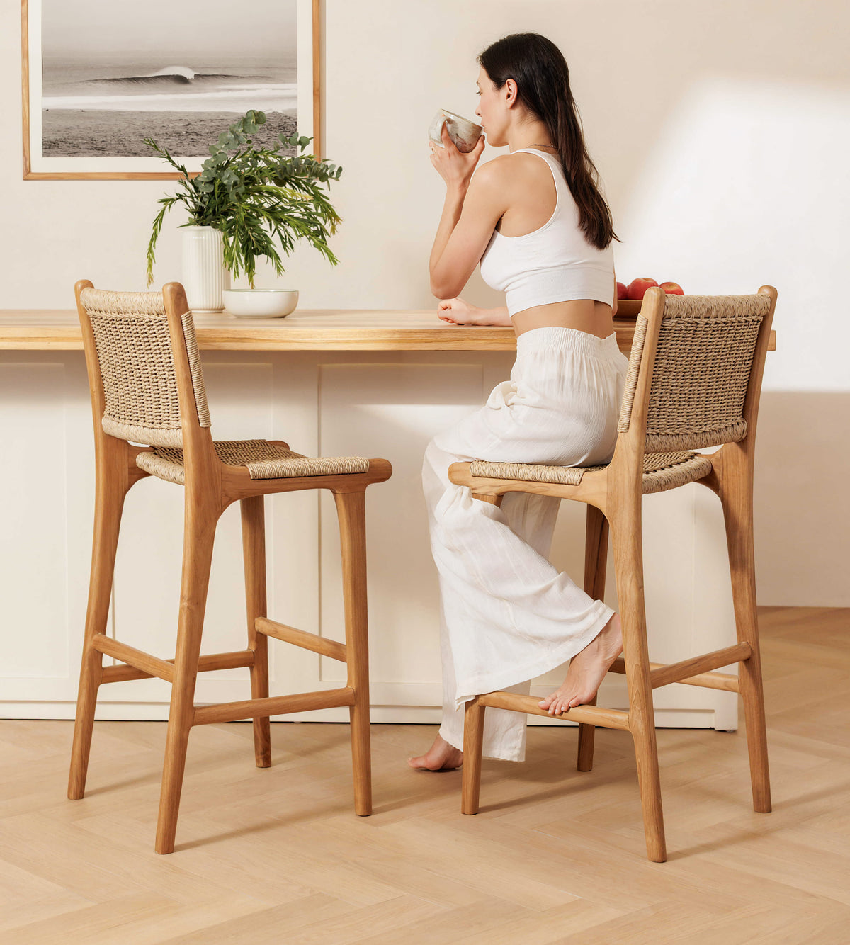 A woman sits barefoot on a wooden bar stool with a woven rattan seat and back, leaning on a light wood counter in a bright, modern interior. Another matching stool stands empty beside her.